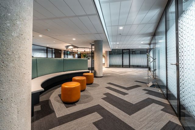 Modern office lobby with curved black bench, orange stools, patterned carpet, and glass walls