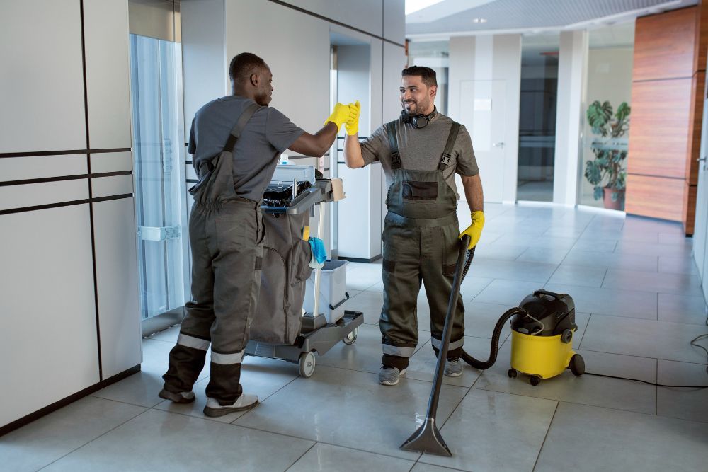 Two technicians in work overalls high-five in a bright hallway beside cleaning equipment.