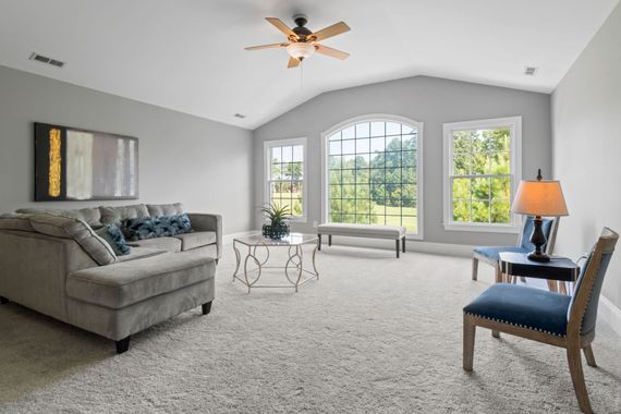 Bright living room with gray sofa, blue chair, large windows, and ceiling fan on carpeted floor