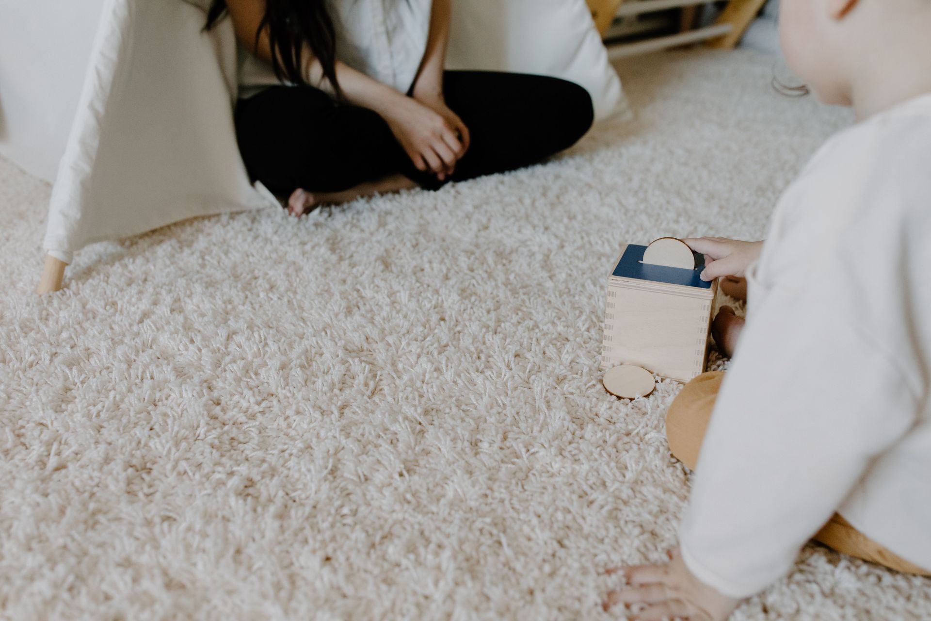Two people sitting on a fluffy white rug, playing with a small wooden toy block in a bright room