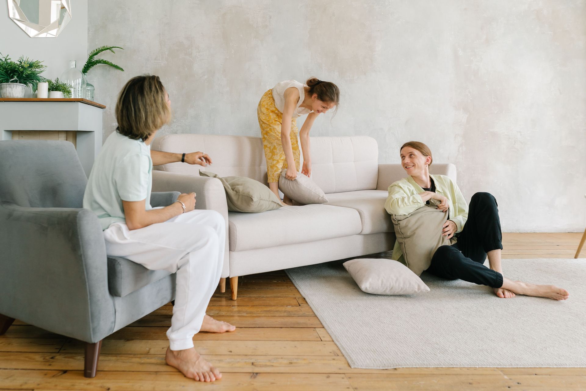 Three people relaxing in a bright living room, chatting on a white sofa and chair with cushions on the floor.