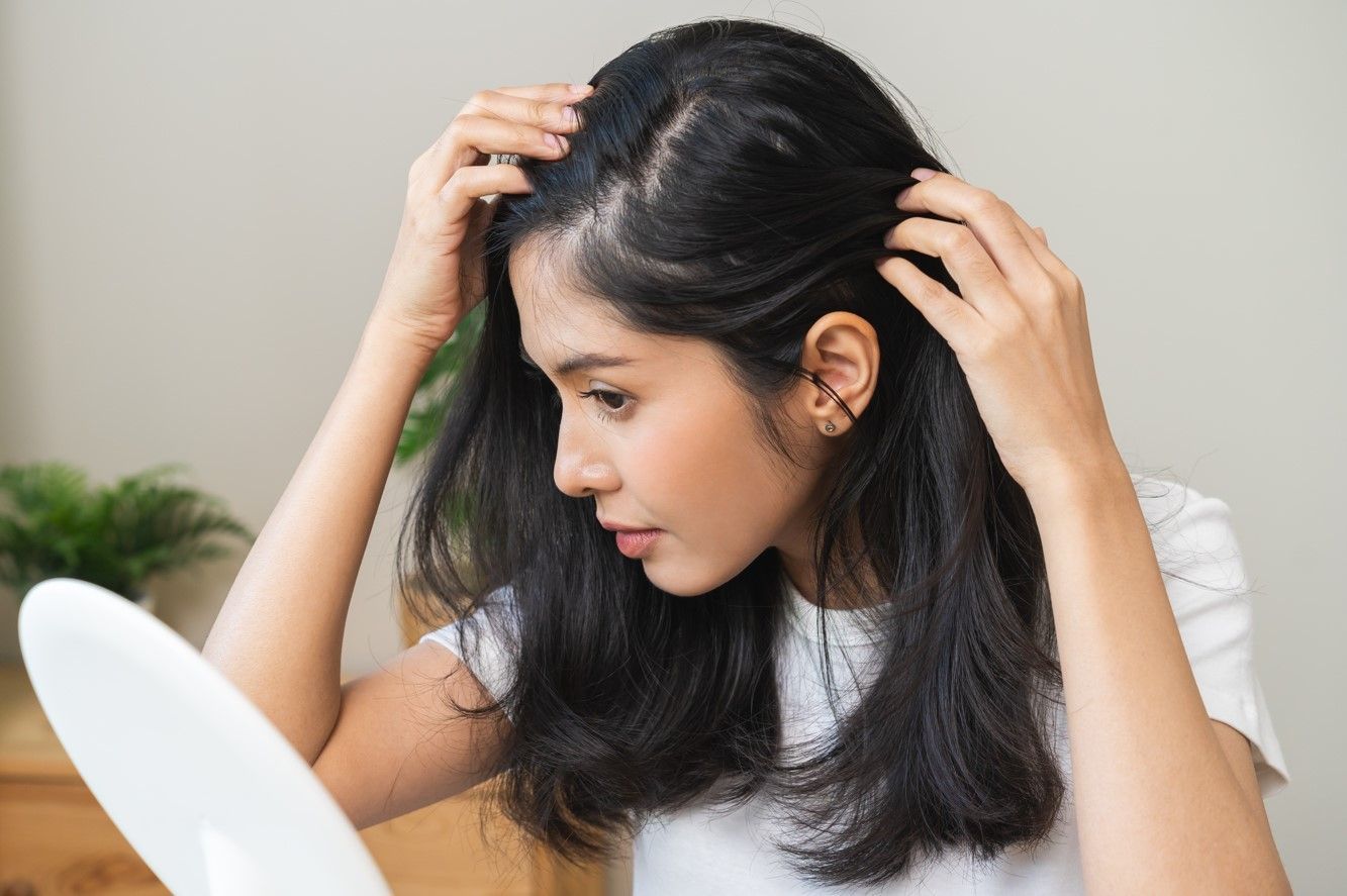 Woman examining her scalp for hair loss, using hands to lift hair. Indoor setting.