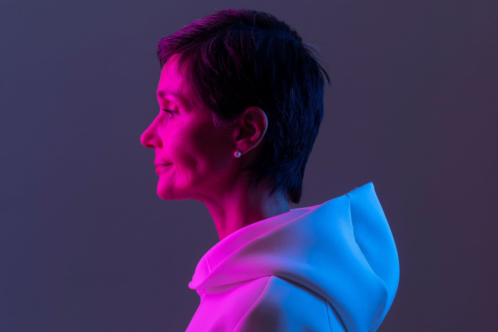 Woman in a white hoodie, side profile. Pink and blue neon lighting.