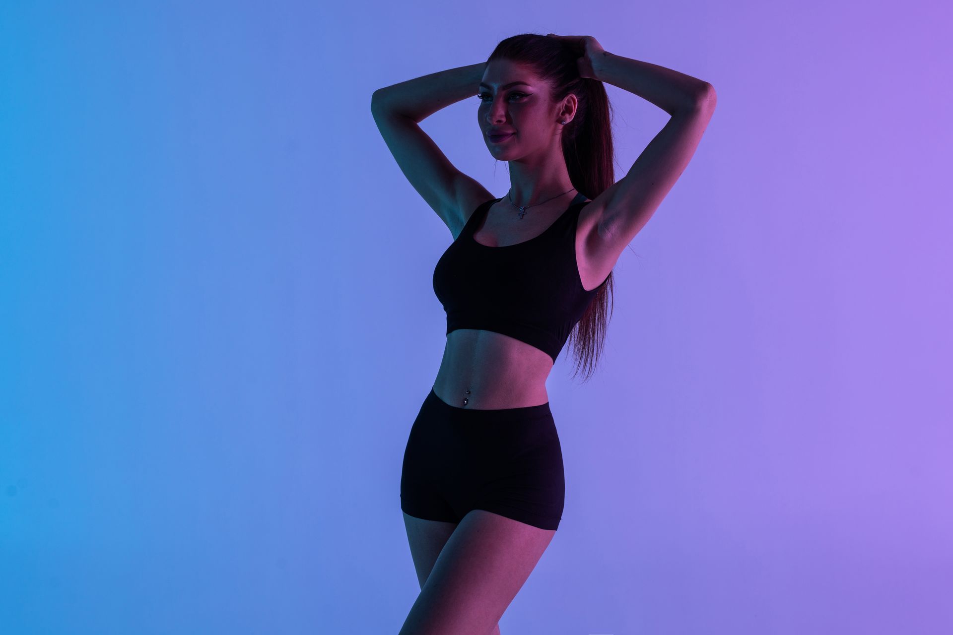 Woman in black workout clothes poses with hands behind head, against a blue and purple gradient background.