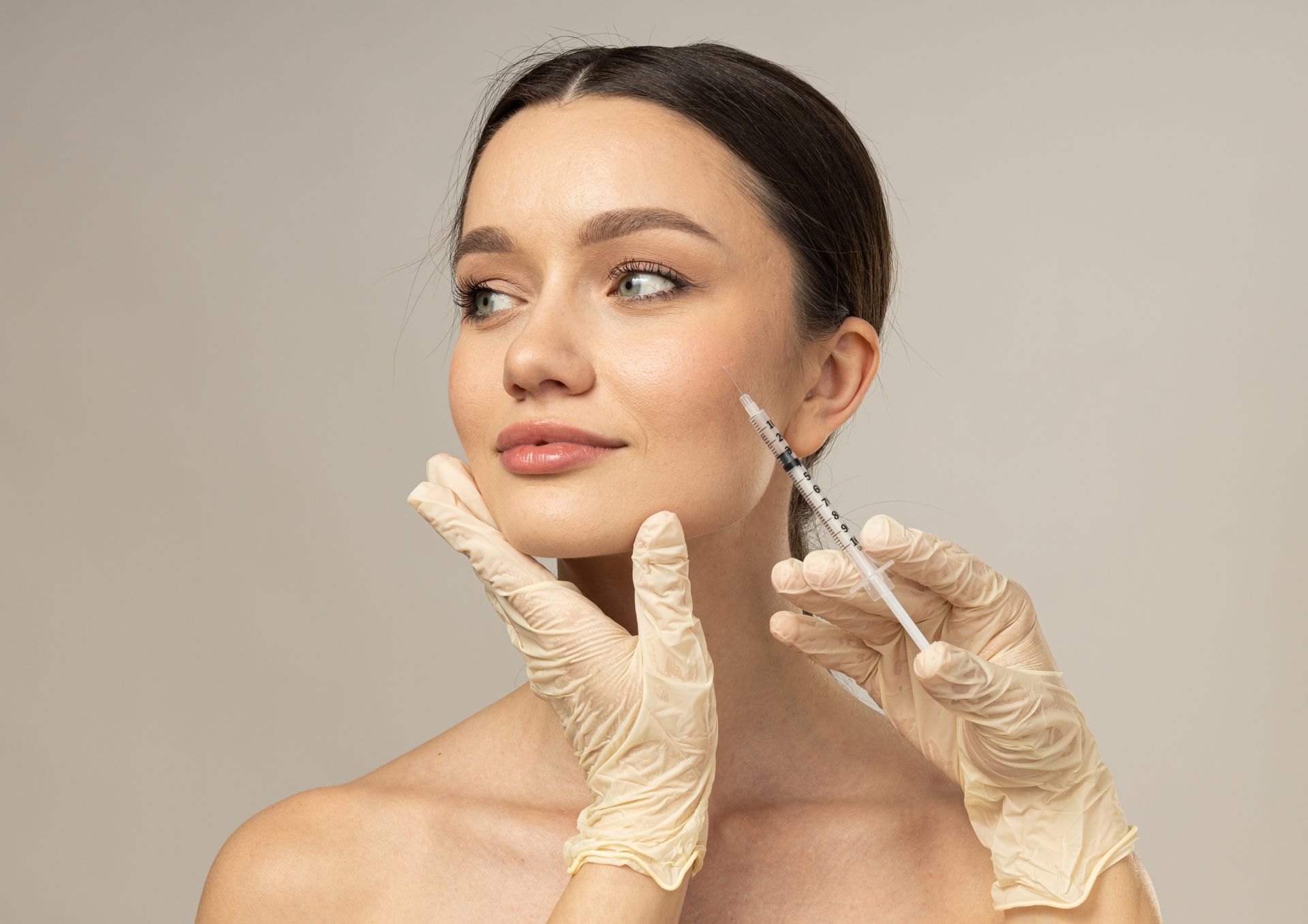 A person receiving an injection in their cheek; gloved hands hold syringe. Beige background.