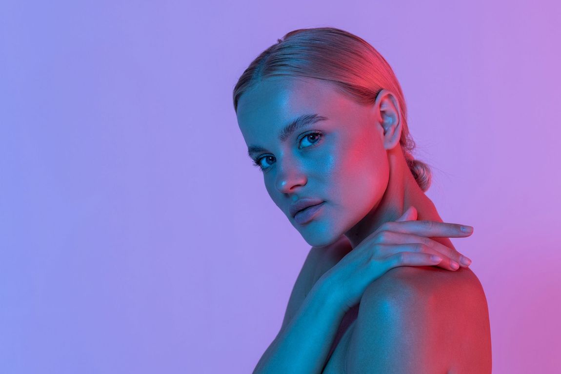 Woman with fair skin, lit with pink and blue light, gazing over shoulder, hand on opposite shoulder.