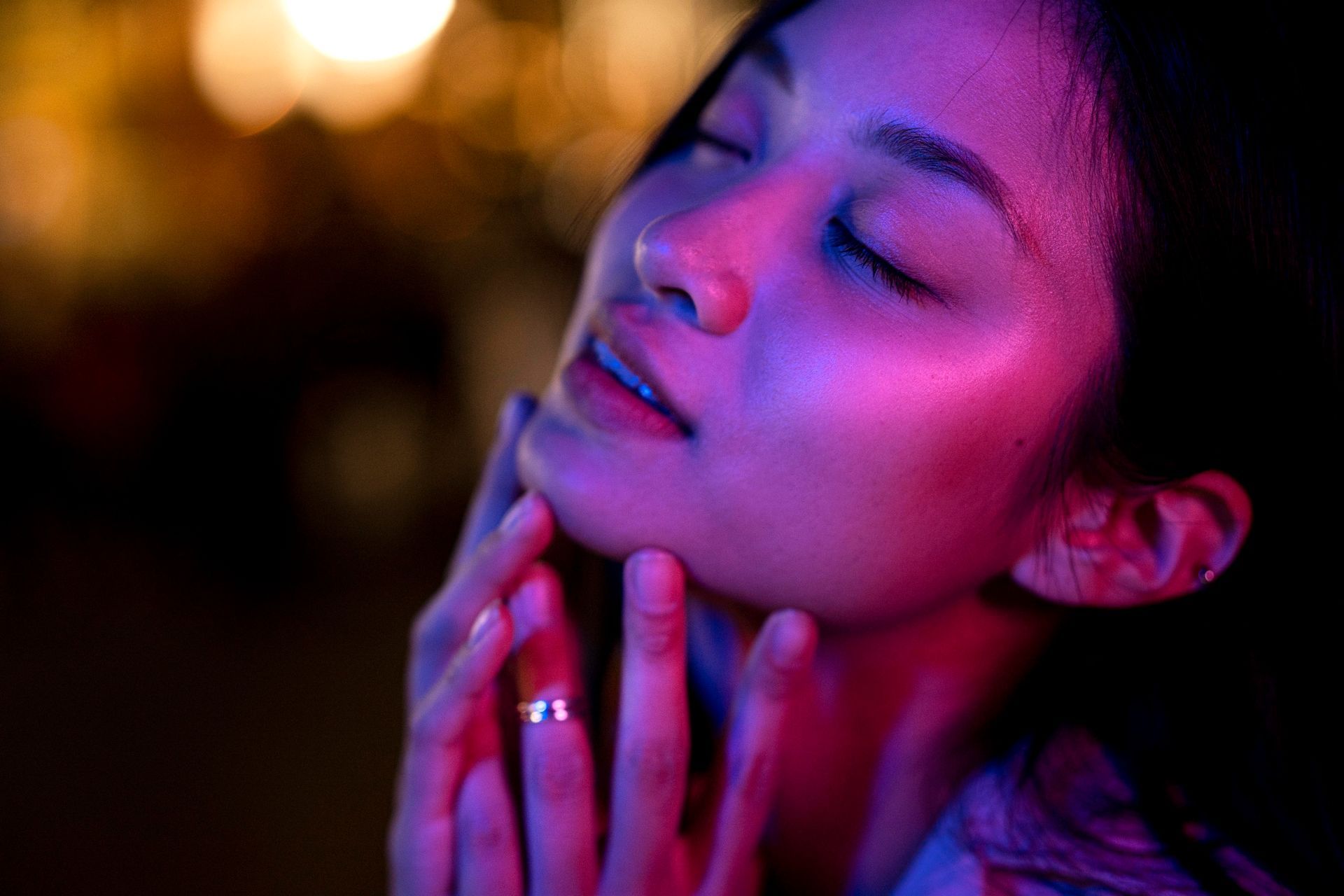 Woman with closed eyes, illuminated by pink and purple light, touching her face.