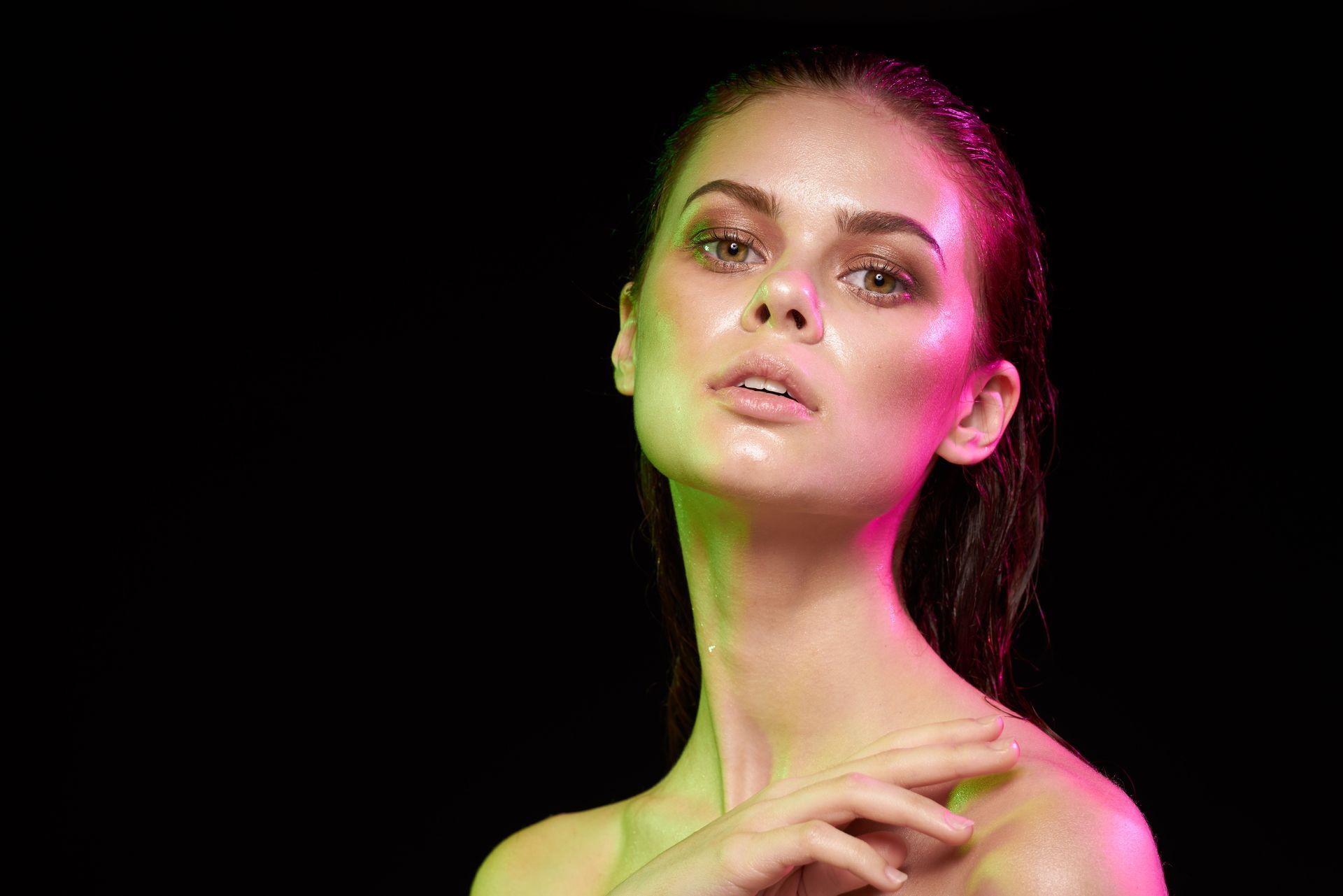 Woman's face illuminated by green and pink lights on a black background, looking upwards.