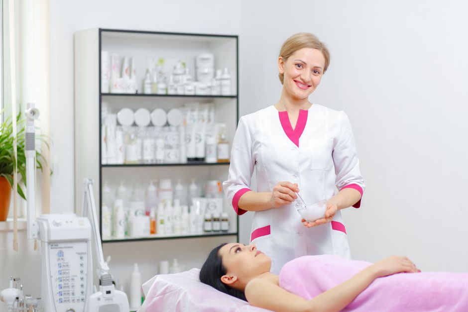 Beautician holding implements, smiling over a patient on a treatment bed in a brightly lit spa room.