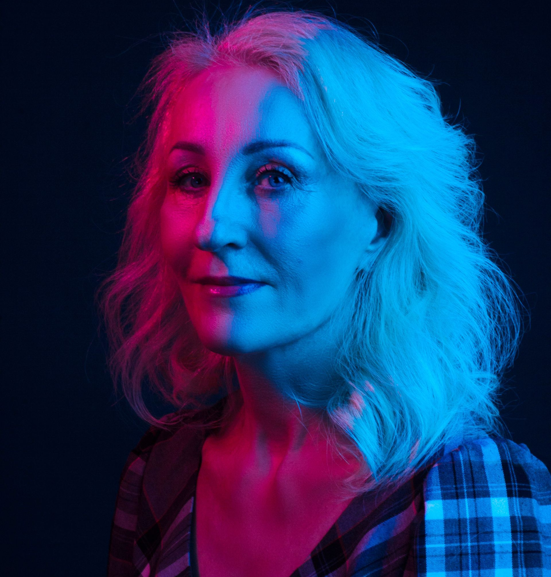 Woman with fair skin, lit by pink and blue light, smiles. She has wavy gray hair and wears a plaid shirt.