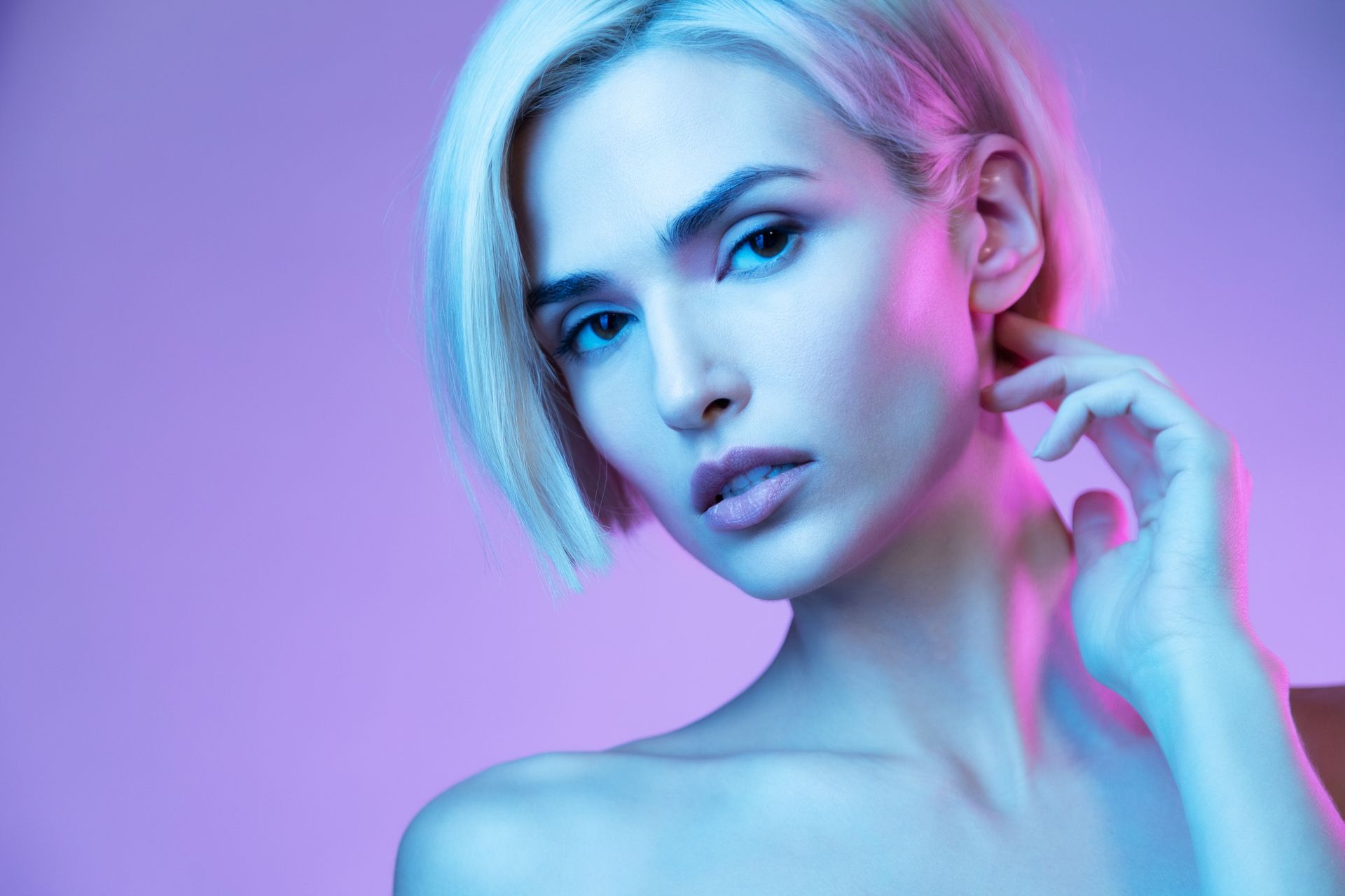 Blonde woman with short hair, illuminated by pink and blue lights, touching her neck, looking at the camera.