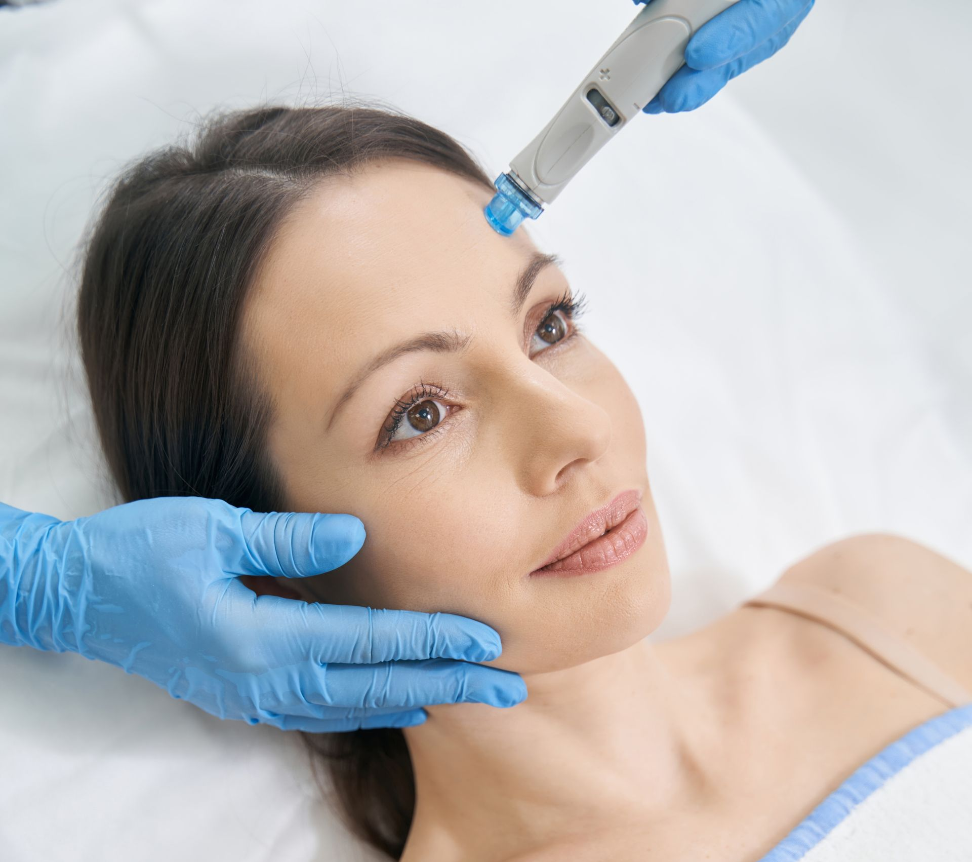 Woman receiving facial treatment; device on forehead, hands in blue gloves.