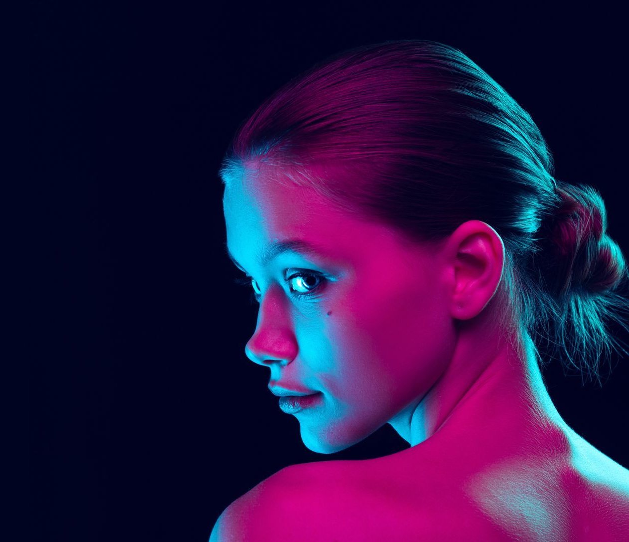 Woman with hair up, lit by pink and blue light, looking over shoulder. Dark background.