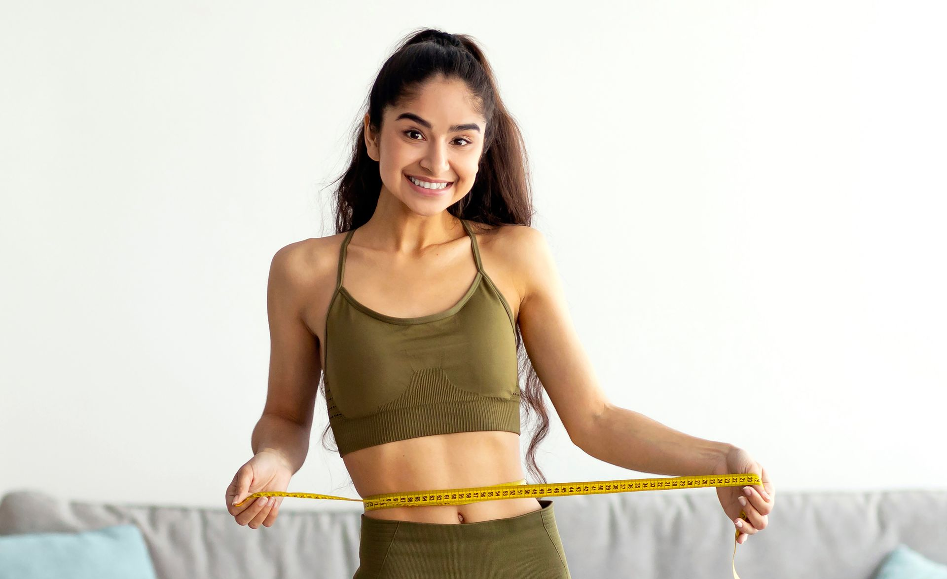 Woman in athletic wear measuring her waist, smiling in a room.