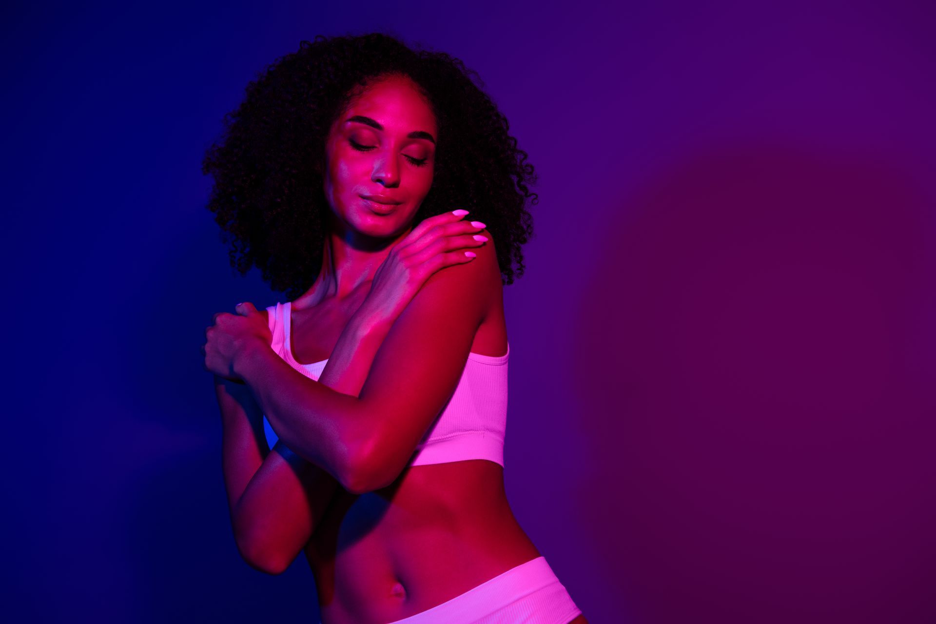 Woman in white underwear, illuminated by pink and blue light, hugs herself, eyes closed.
