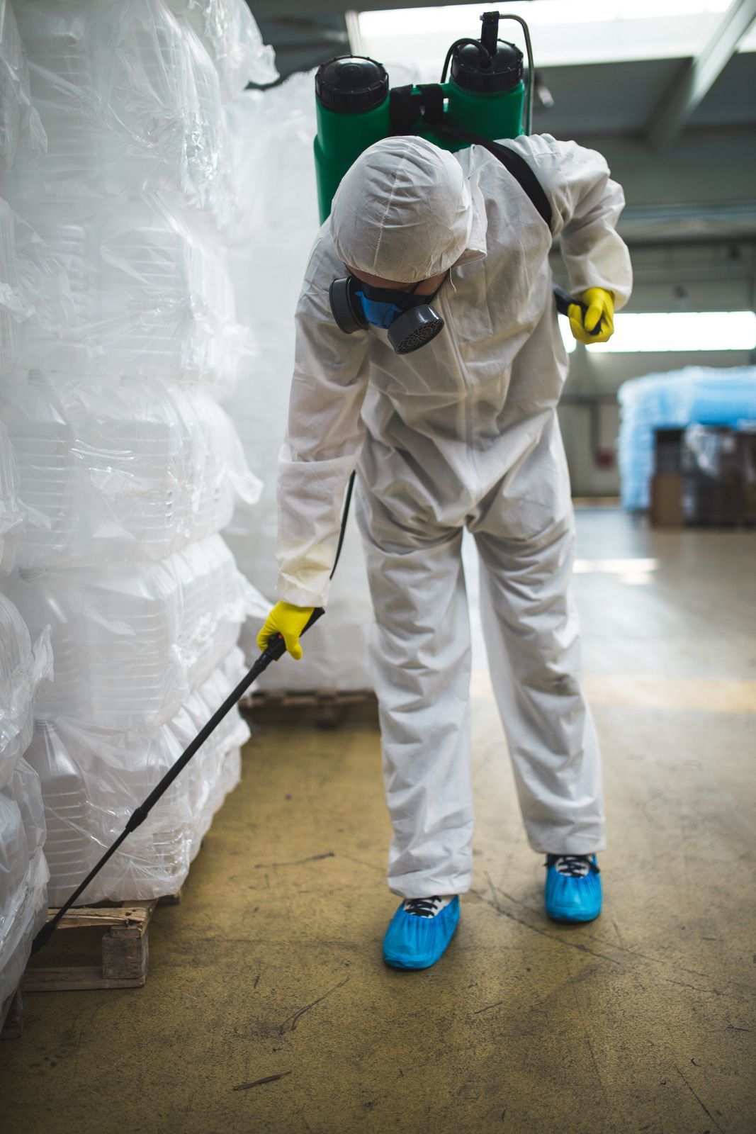 A worker in a white hazmat suit, mask, and yellow gloves uses a green sprayer to disinfect a warehouse floor.