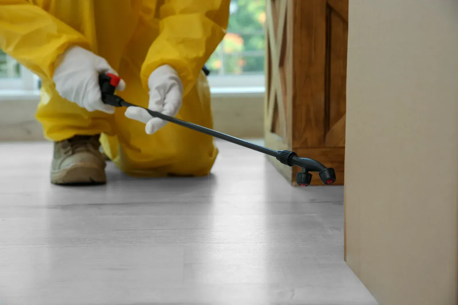 A person in a yellow protective suit and white gloves sprays pesticide along a wooden baseboard indoors.