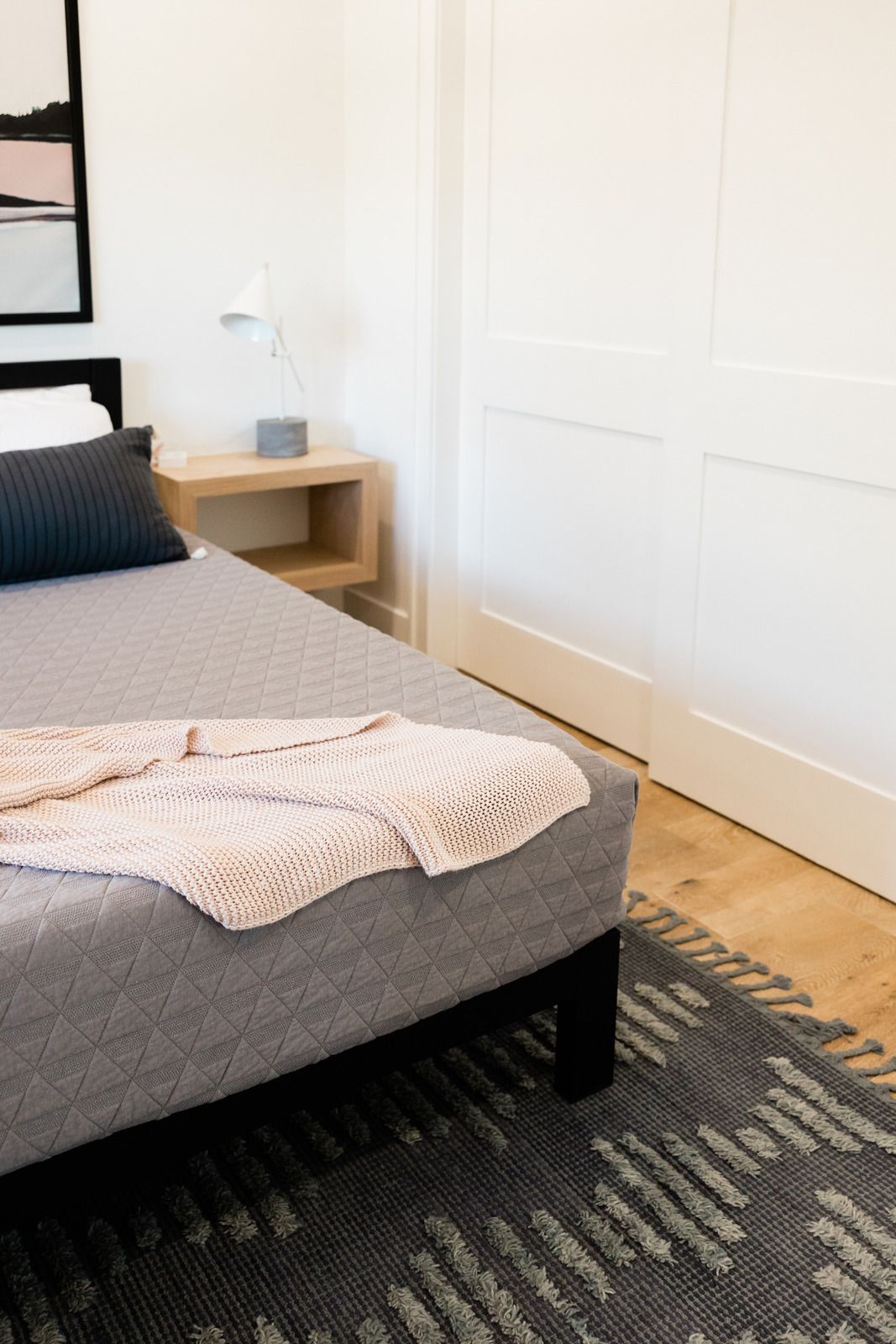 A modern bedroom featuring a gray bed with a textured blanket, a wooden nightstand, a desk lamp, and a patterned rug.