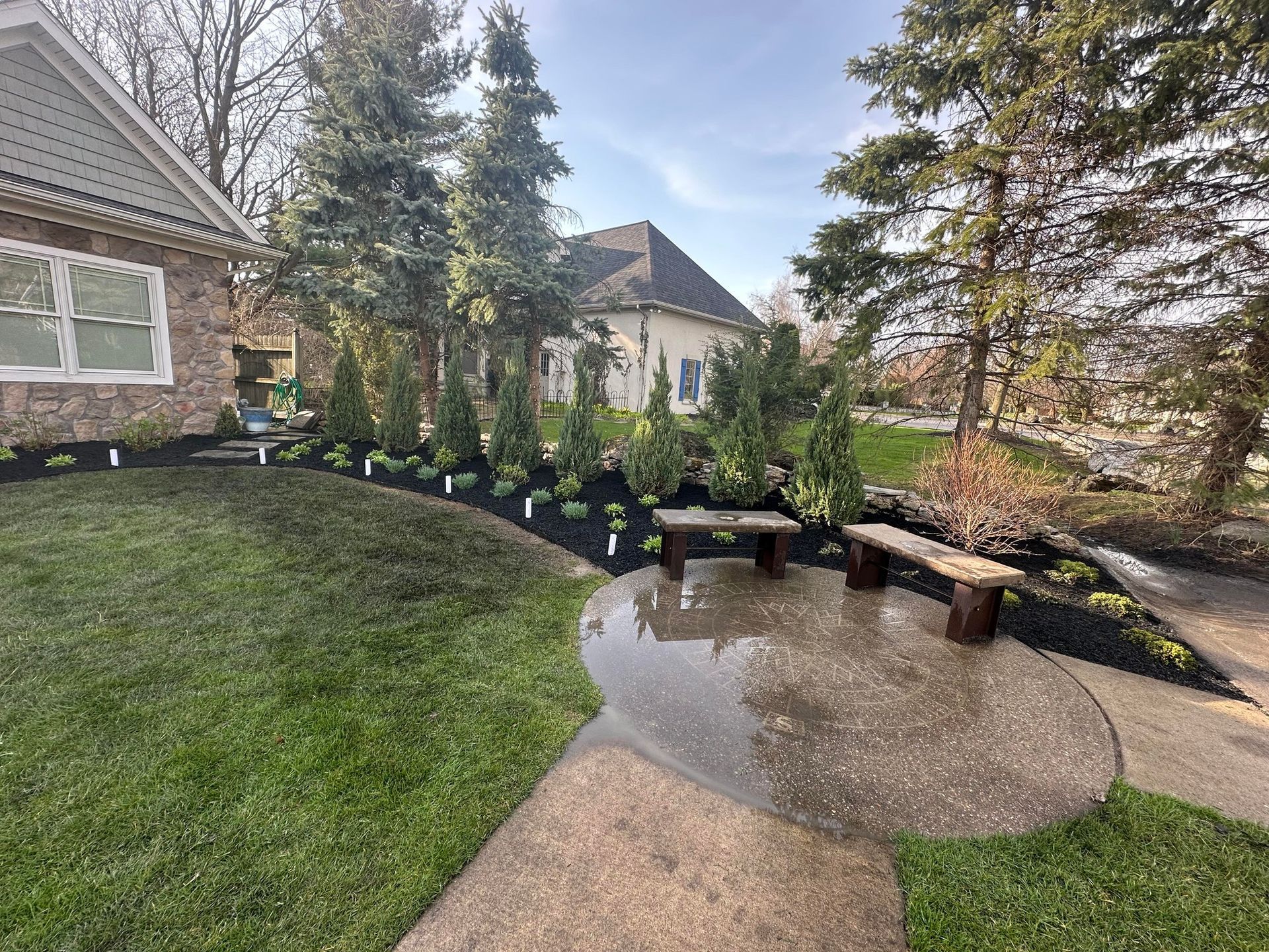 Lush green lawn slopes to a stone patio with benches. Mulched flowerbeds with evergreen trees flank a house. Landscaping 