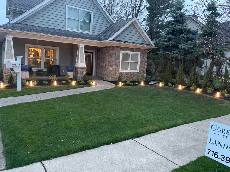 A well-manicured lawn with lighted landscaping leading to a house with gray siding and stone accents.