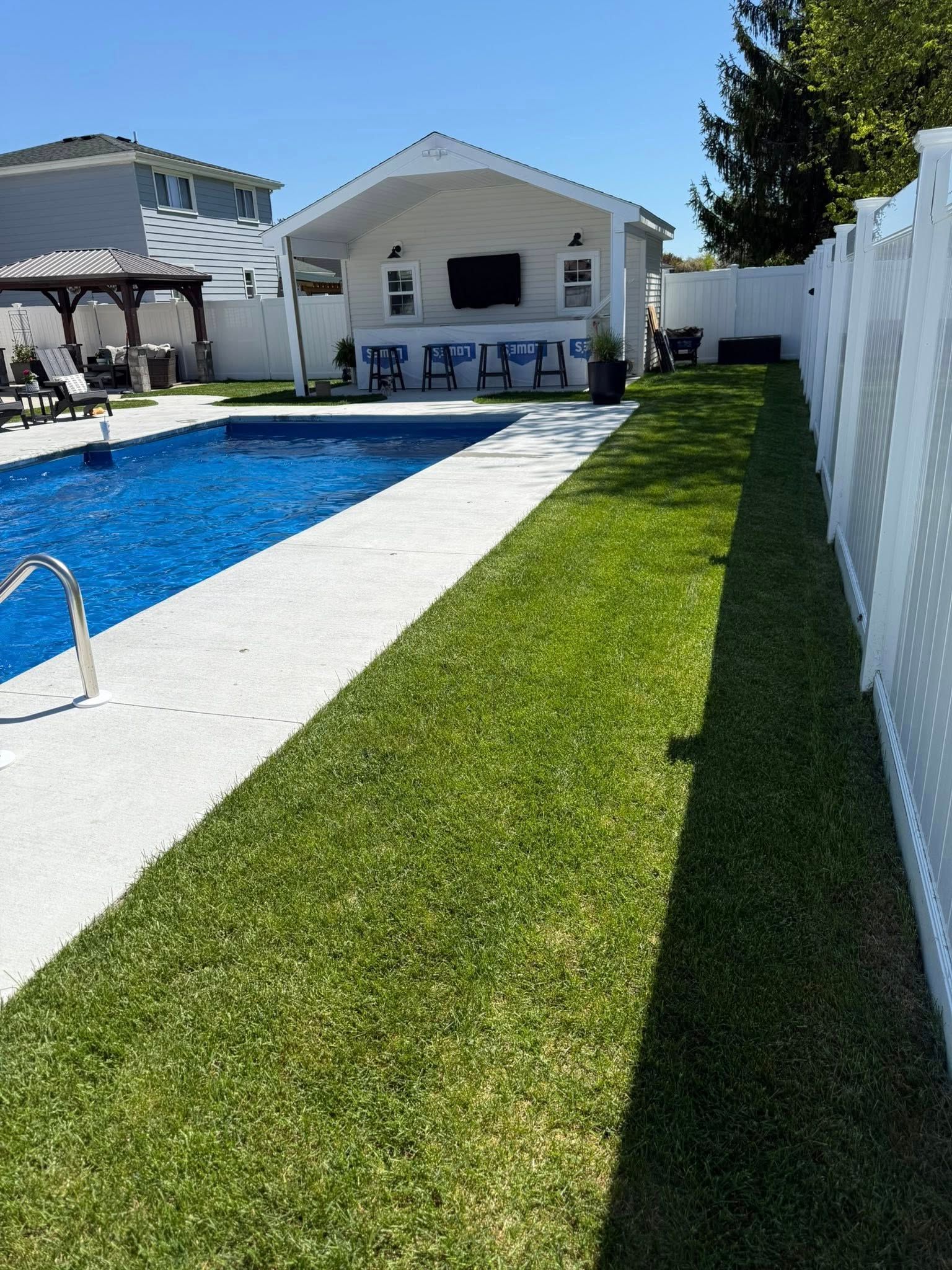 A backyard with a pool, bar area, and white fence; green lawn and blue sky. Sod INstallation