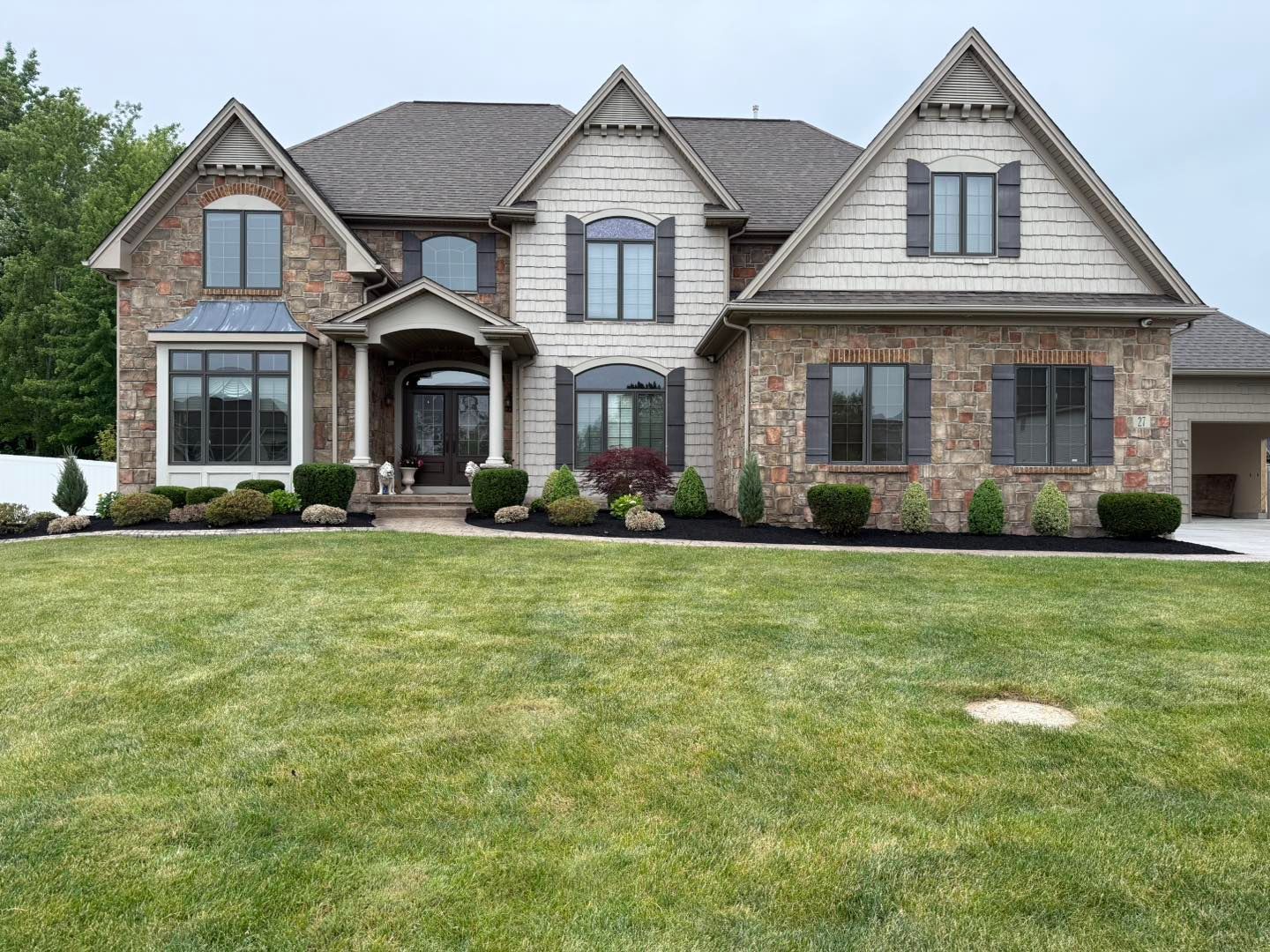 Two-story house with stone and tan siding, brown roof, dark shutters, and a well-manicured lawn.