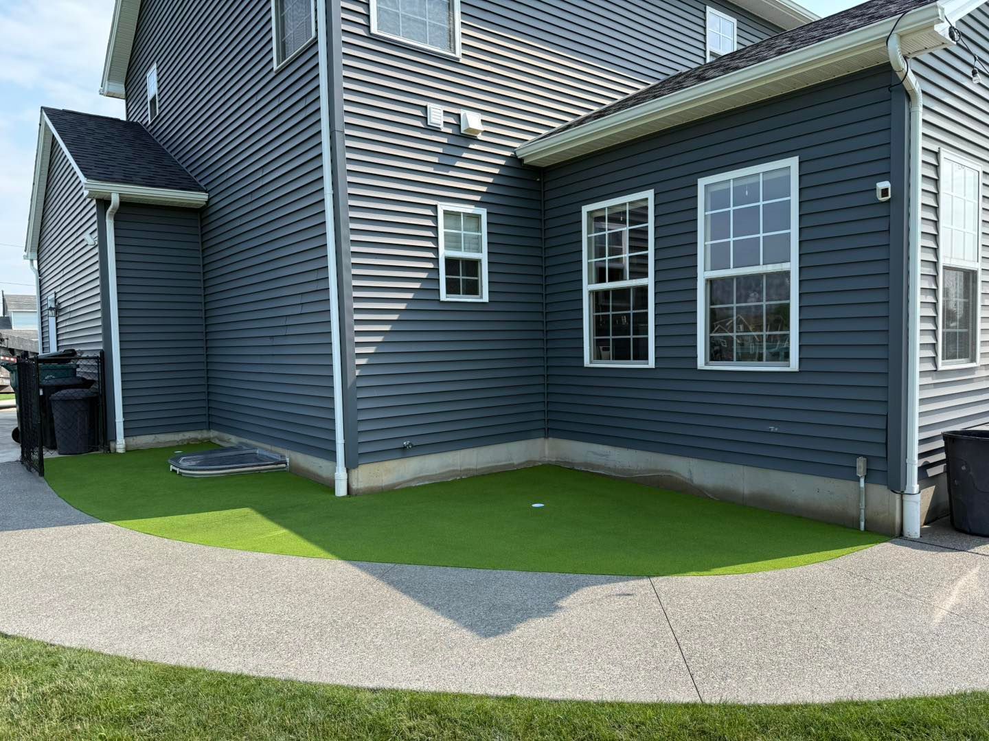 Backyard with a dark blue house, green artificial turf putting area, and concrete walkway. Turf Installation