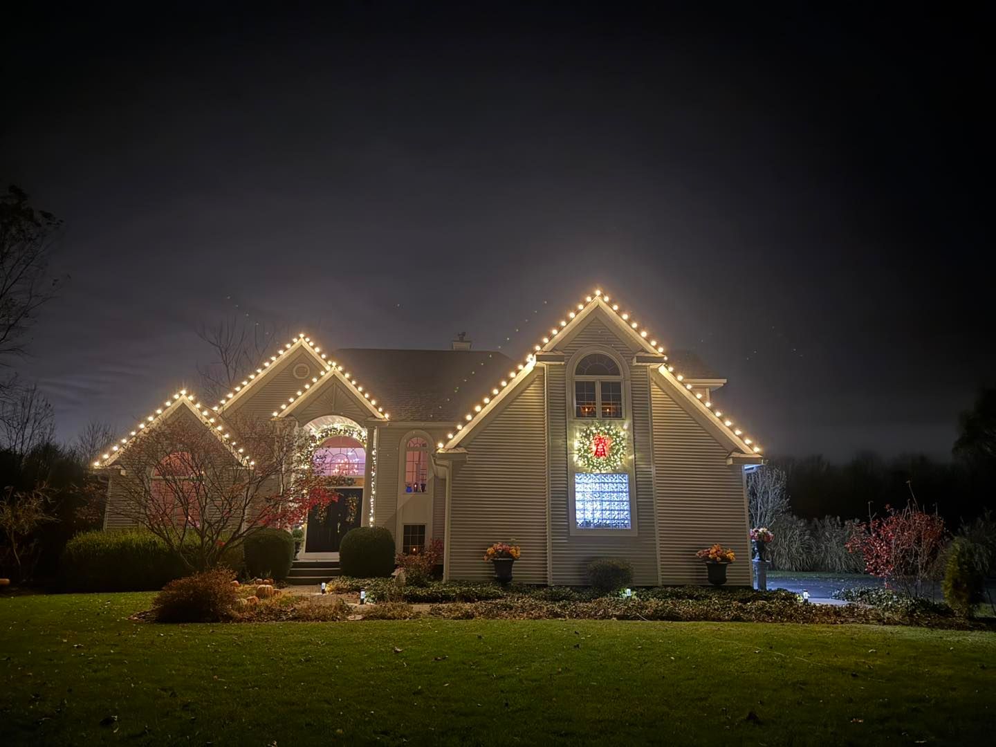 House at night decorated with warm white Christmas lights on roof and wreaths, with lawn in front.