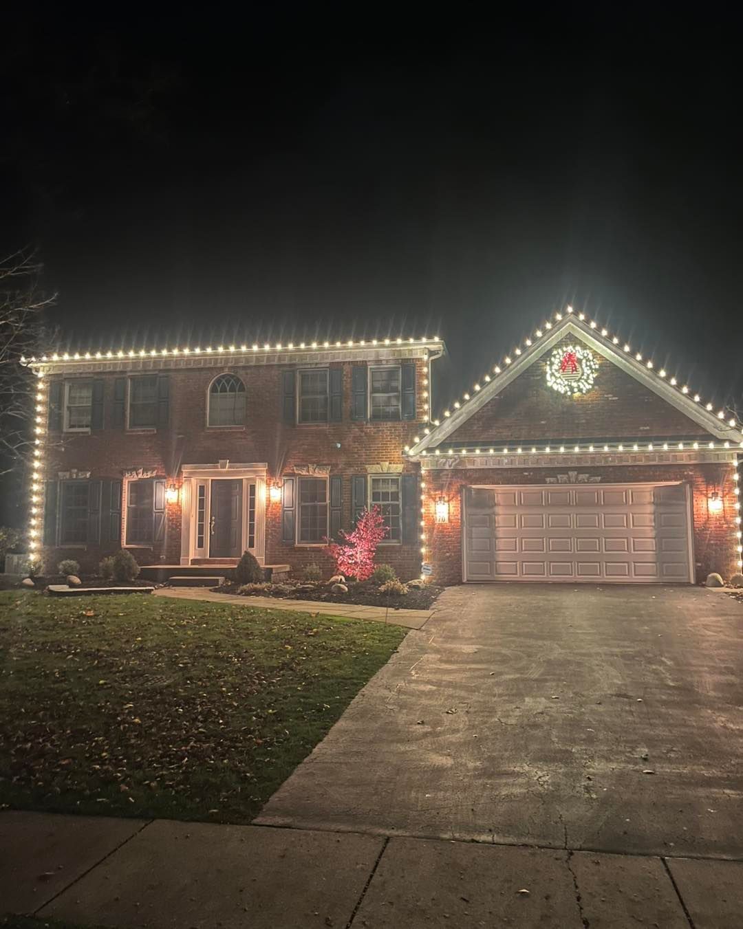 Two-story brick house at night, decorated with white Christmas lights along rooflines and a wreath.
