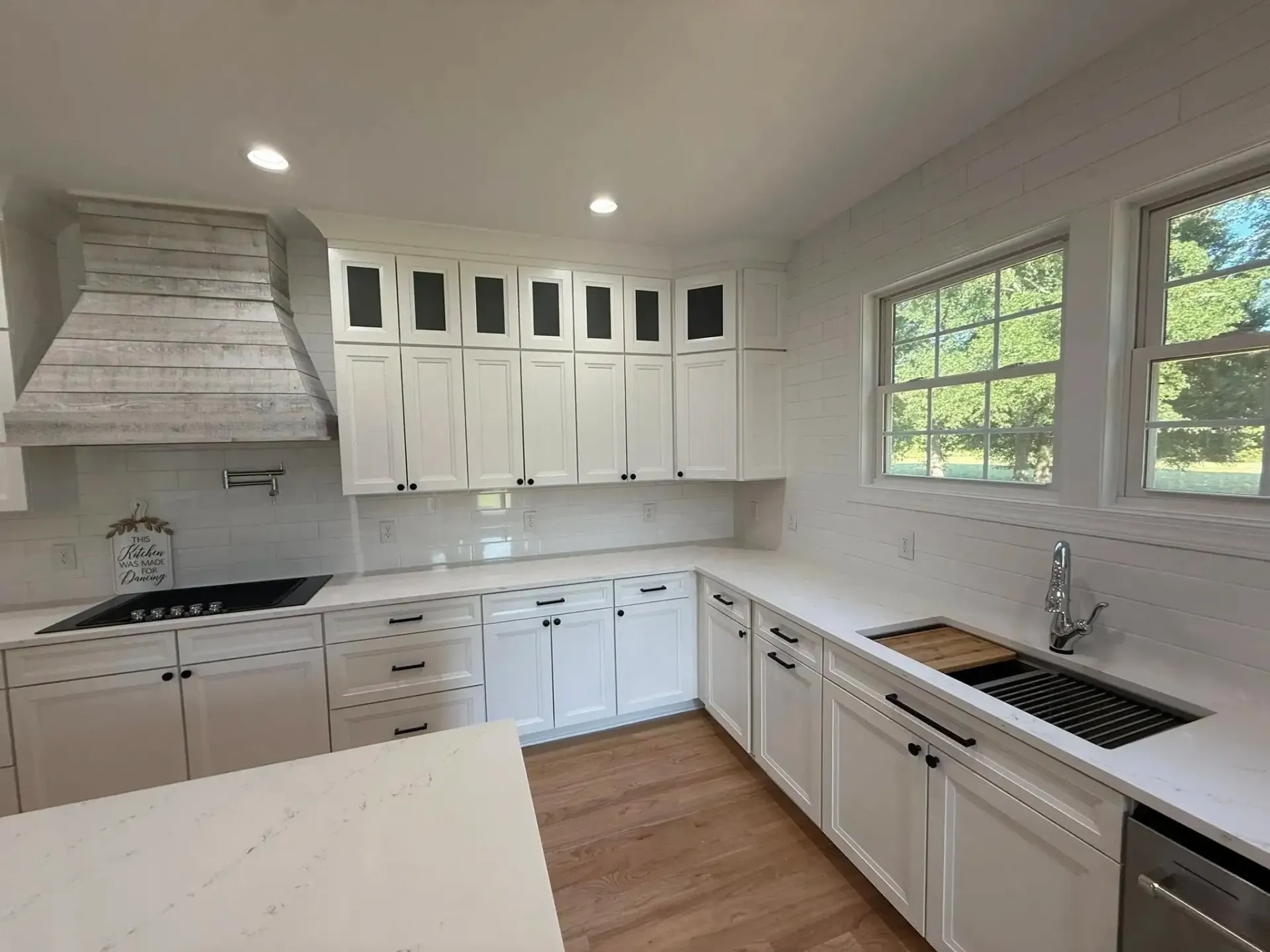 Bright white kitchen with cabinets, black countertops, farmhouse sink, and two large windows overlooking greenery