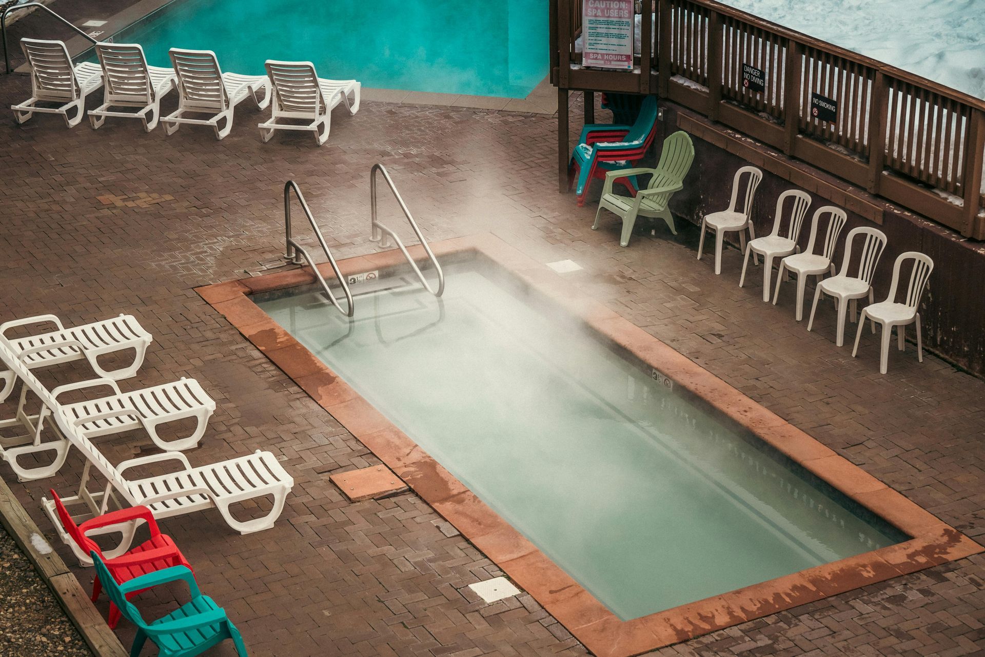 Outdoor hot tub and pool with lounge chairs and steam rising beside a wooden deck and railing