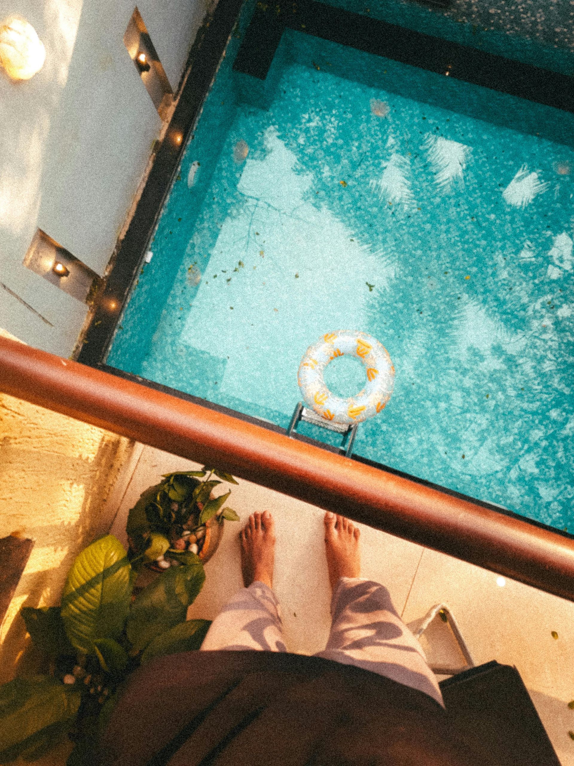 Overhead view of feet by a railing above bright turquoise pool water with a floating ring