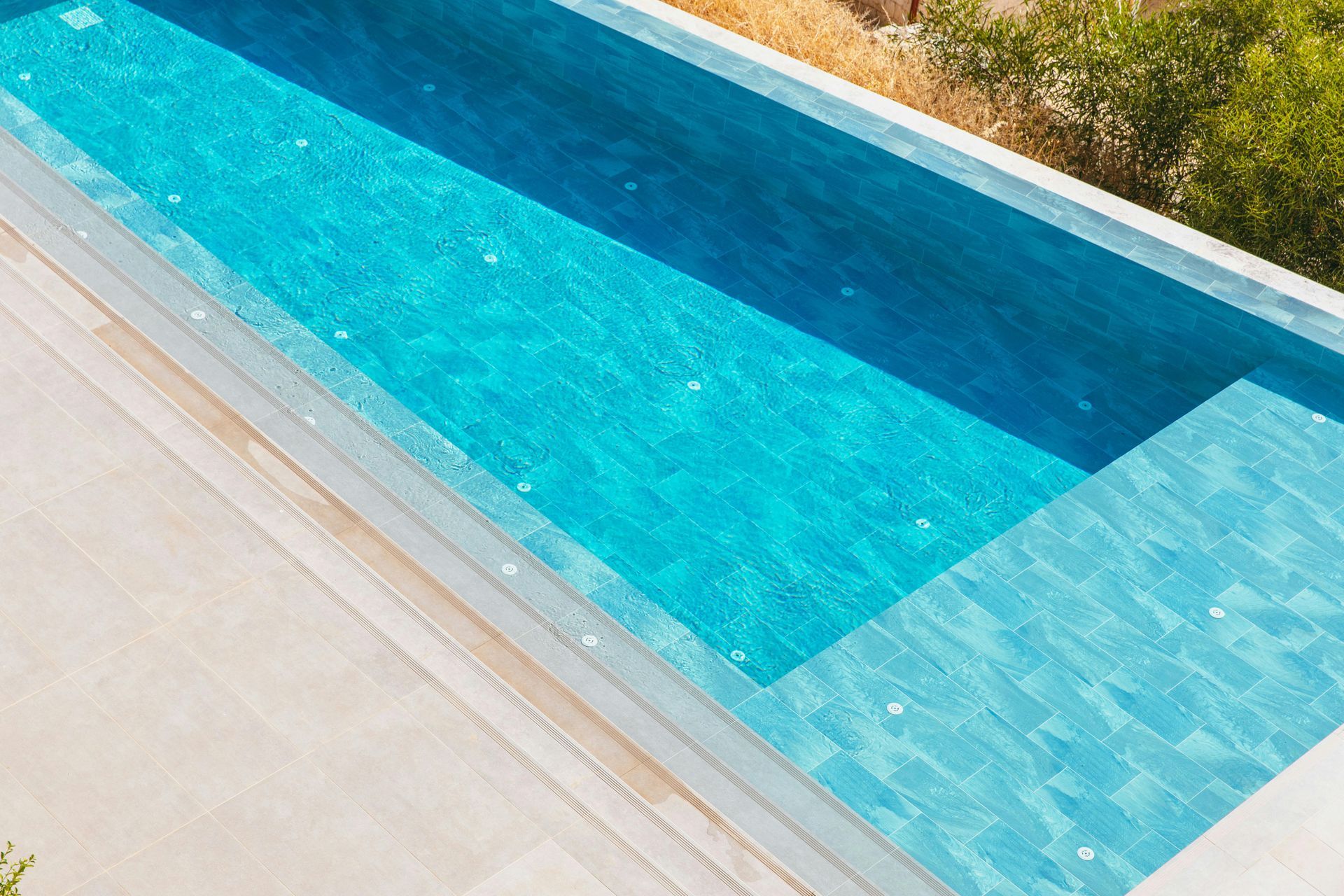 Blue rectangular swimming pool with tiled water and a light stone deck beside it.