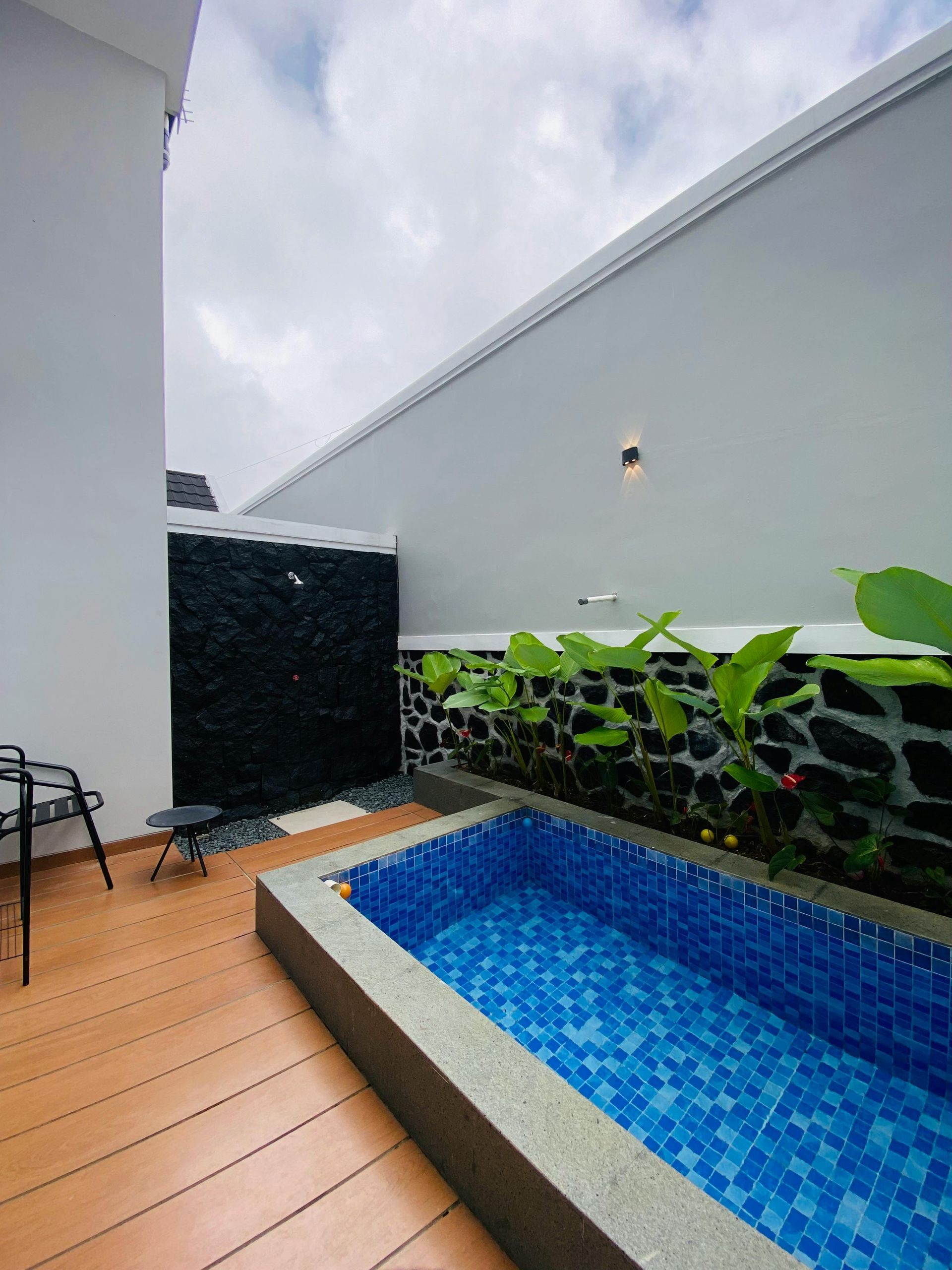 Small rooftop patio with a blue-tiled pool, wooden deck, gray walls, and potted plants under an overcast sky