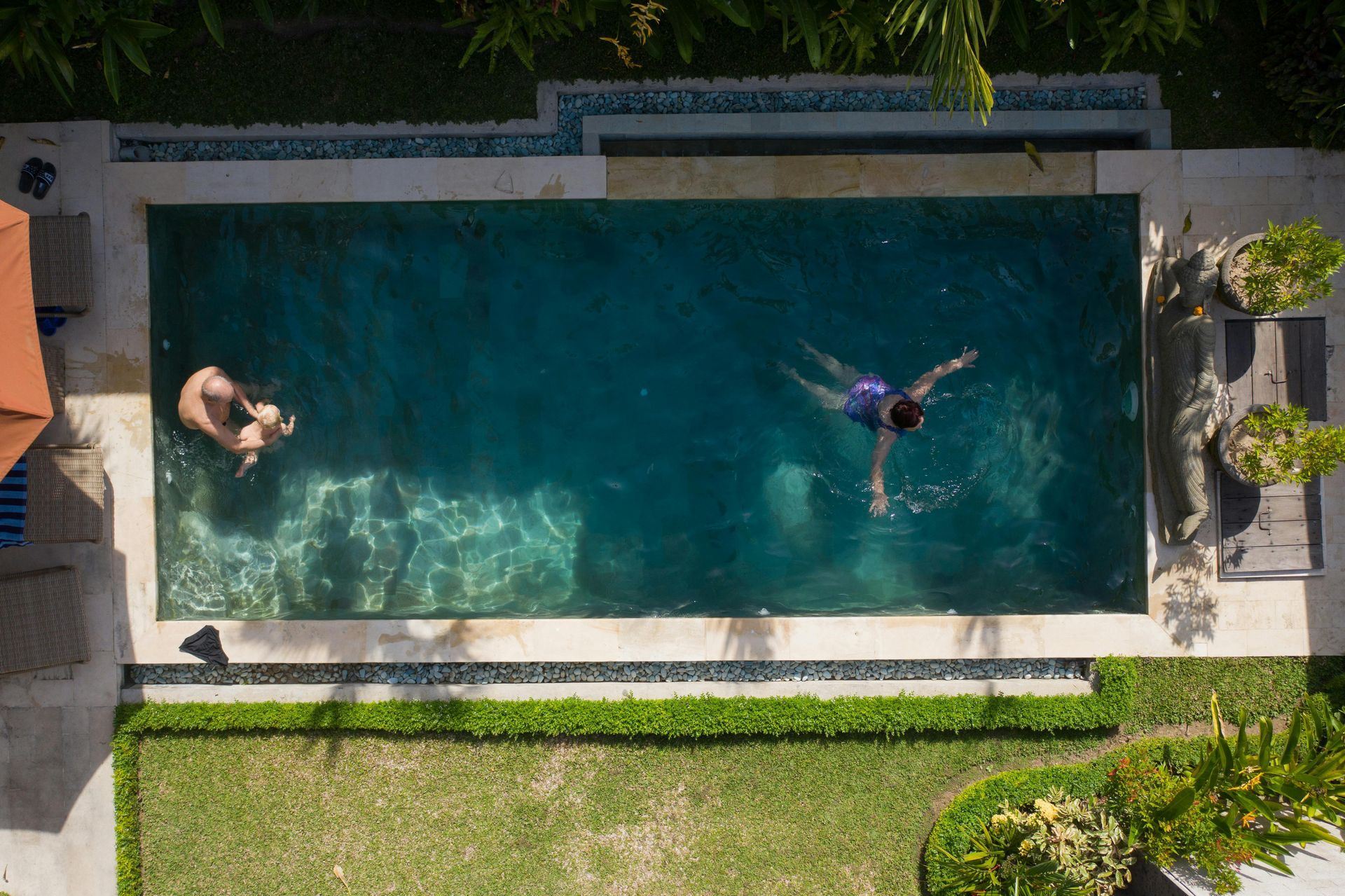 Aerial view of two people swimming in a rectangular backyard pool surrounded by stone patio and grass.