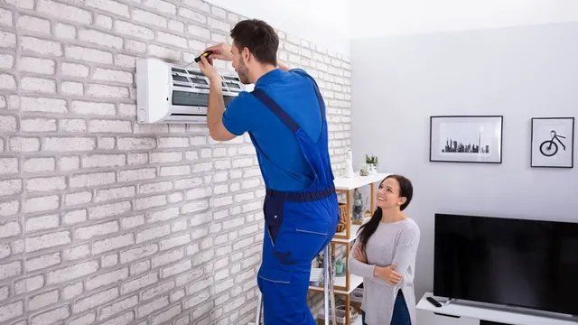 A repairman in blue overalls works on an air conditioner while a woman watches, both in a living room.