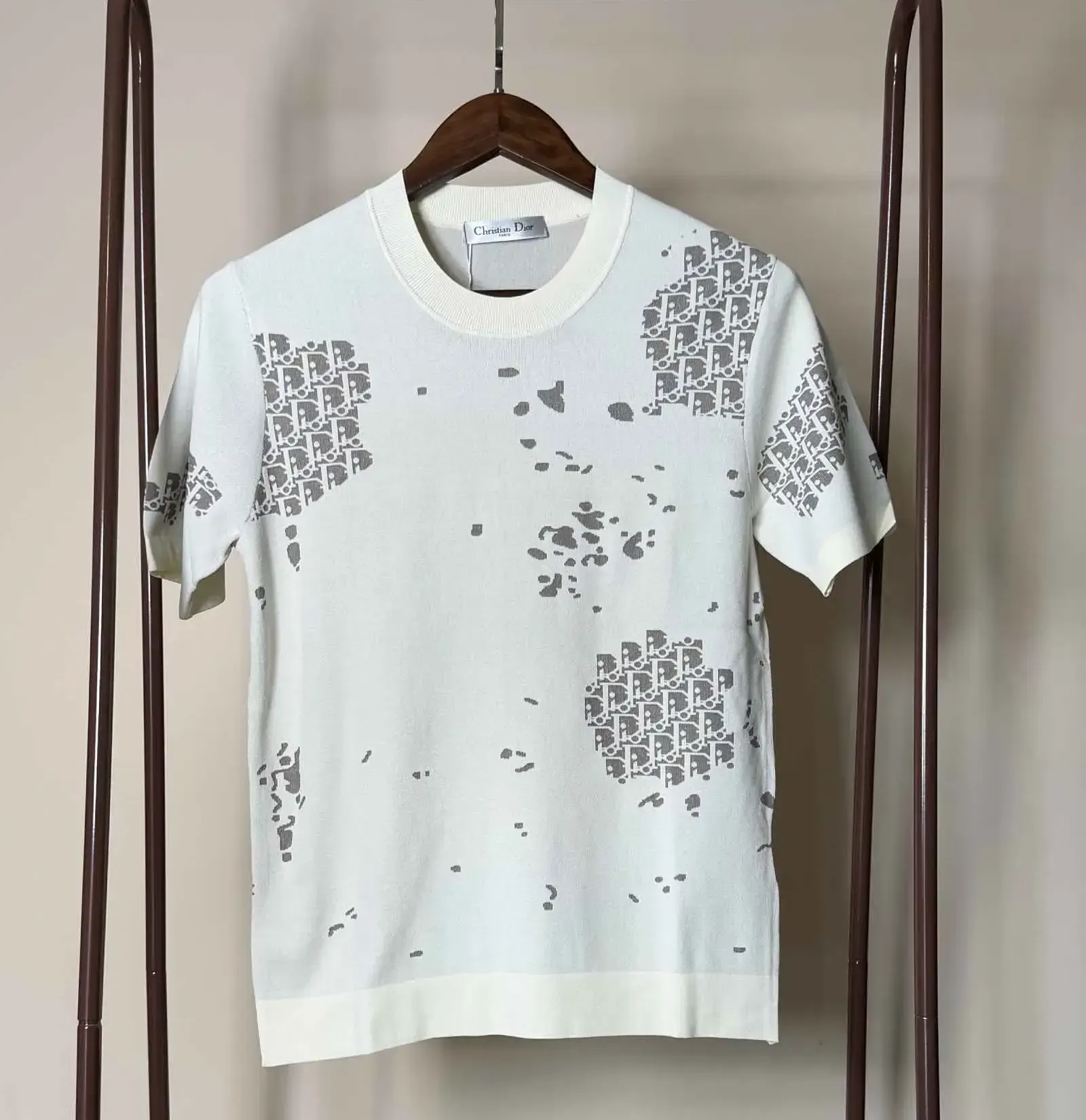 White knit t-shirt with gray logo pattern hanging on a wooden hanger on a brown rack.