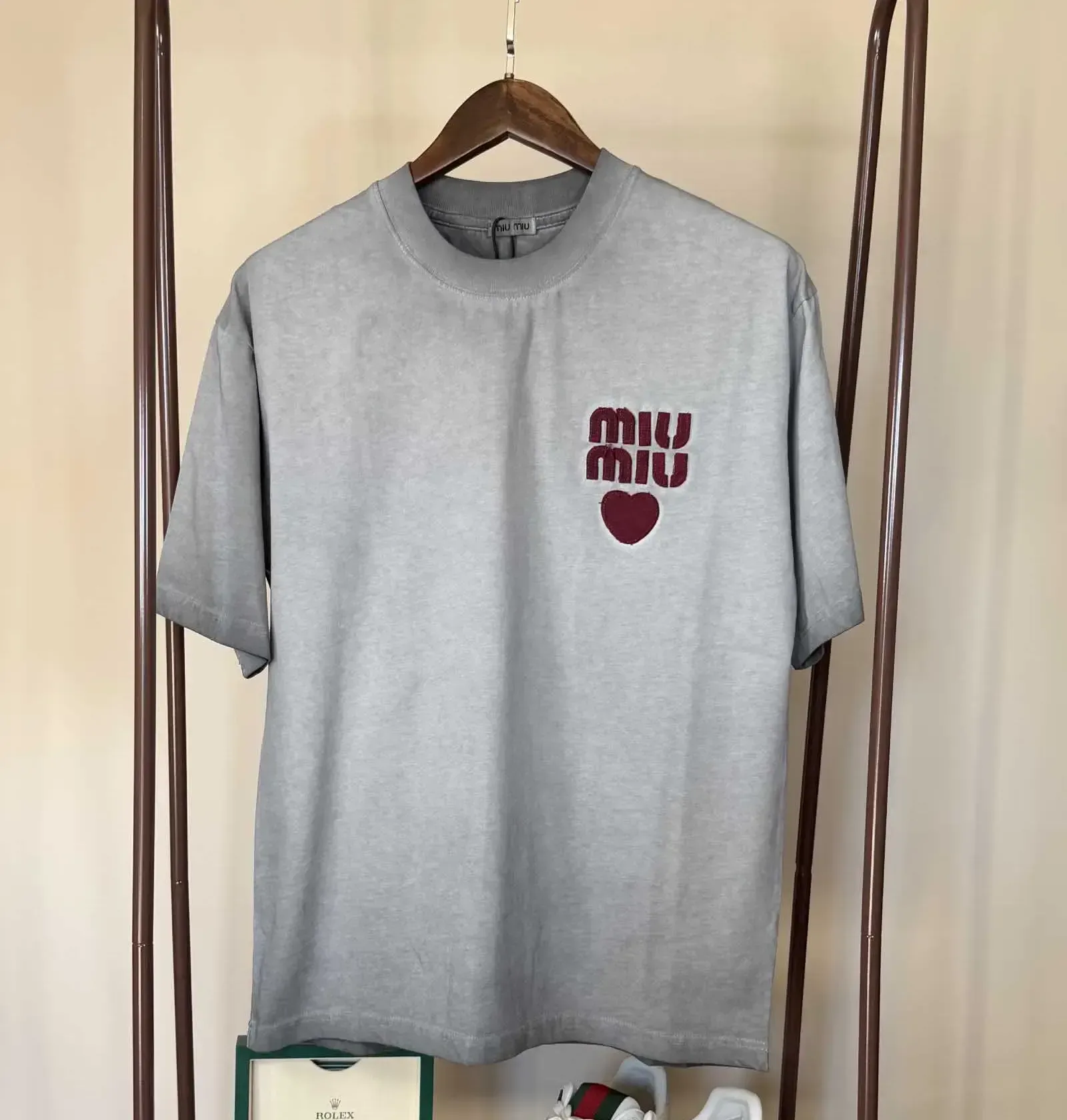 Gray Miu Miu t-shirt with logo and heart graphic hanging on a wooden hanger on a clothes rack.