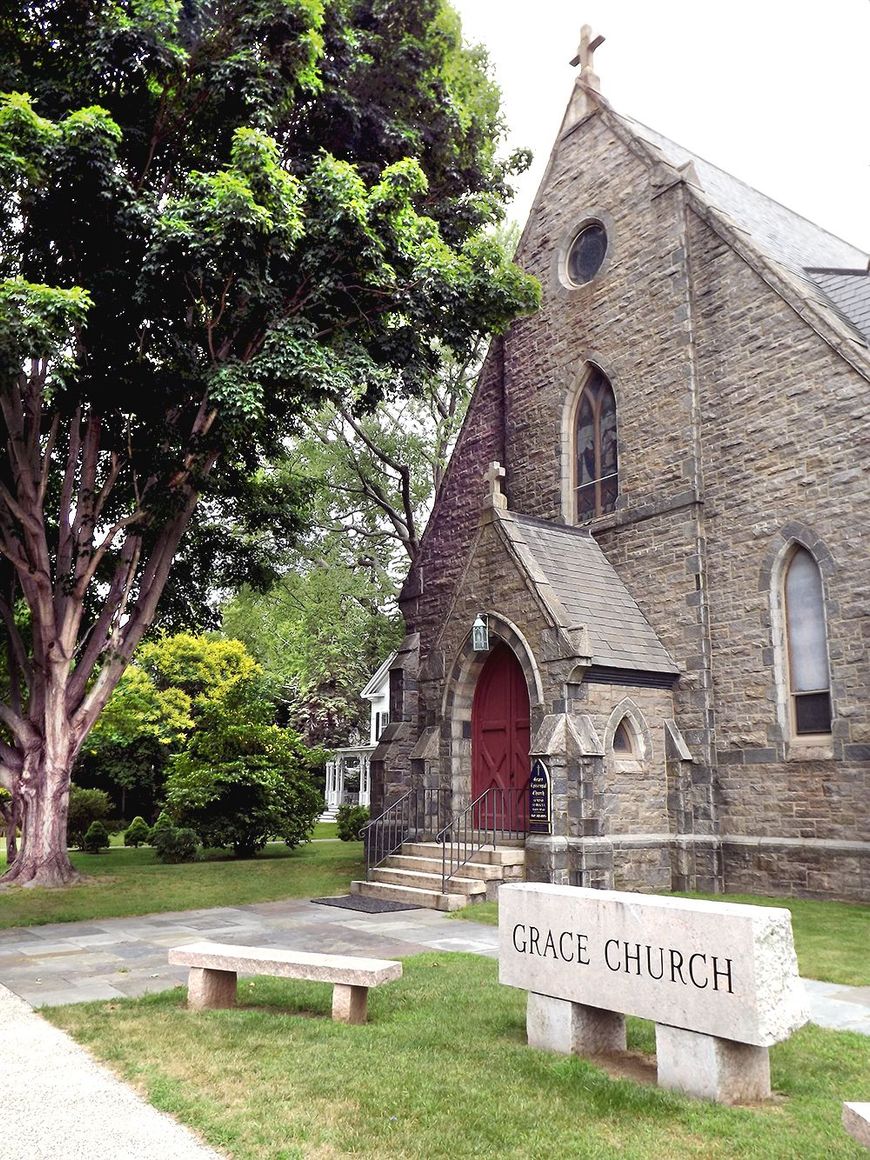 Stone church front entrance with red door, beside a large tree and stone bench near an engraved stone sign, on a grassy lawn