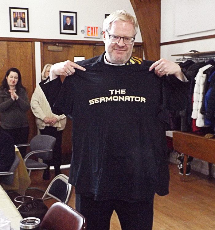 Pastor Charlie holding up a black T-shirt reading “THE SERMONATOR” in the Parish Hall with surrounding chairs and coats