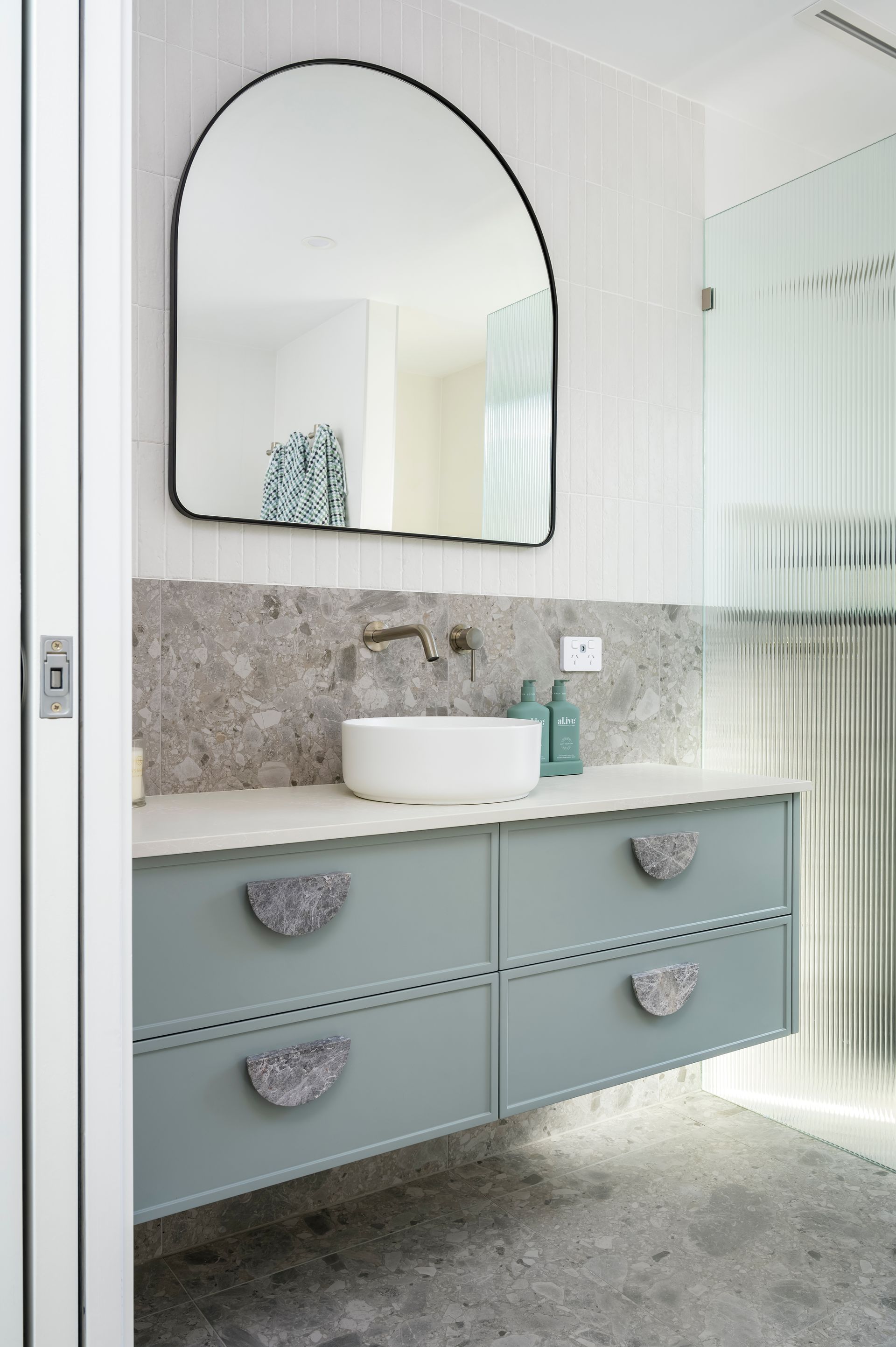 A bathroom vanity with an overmount white round sink, black framed arched mirror, and blue polyurethane drawers in a thin shaker profile with a grey marble half-moon handle. The shower features a frosted fluted glass freestanding screen.