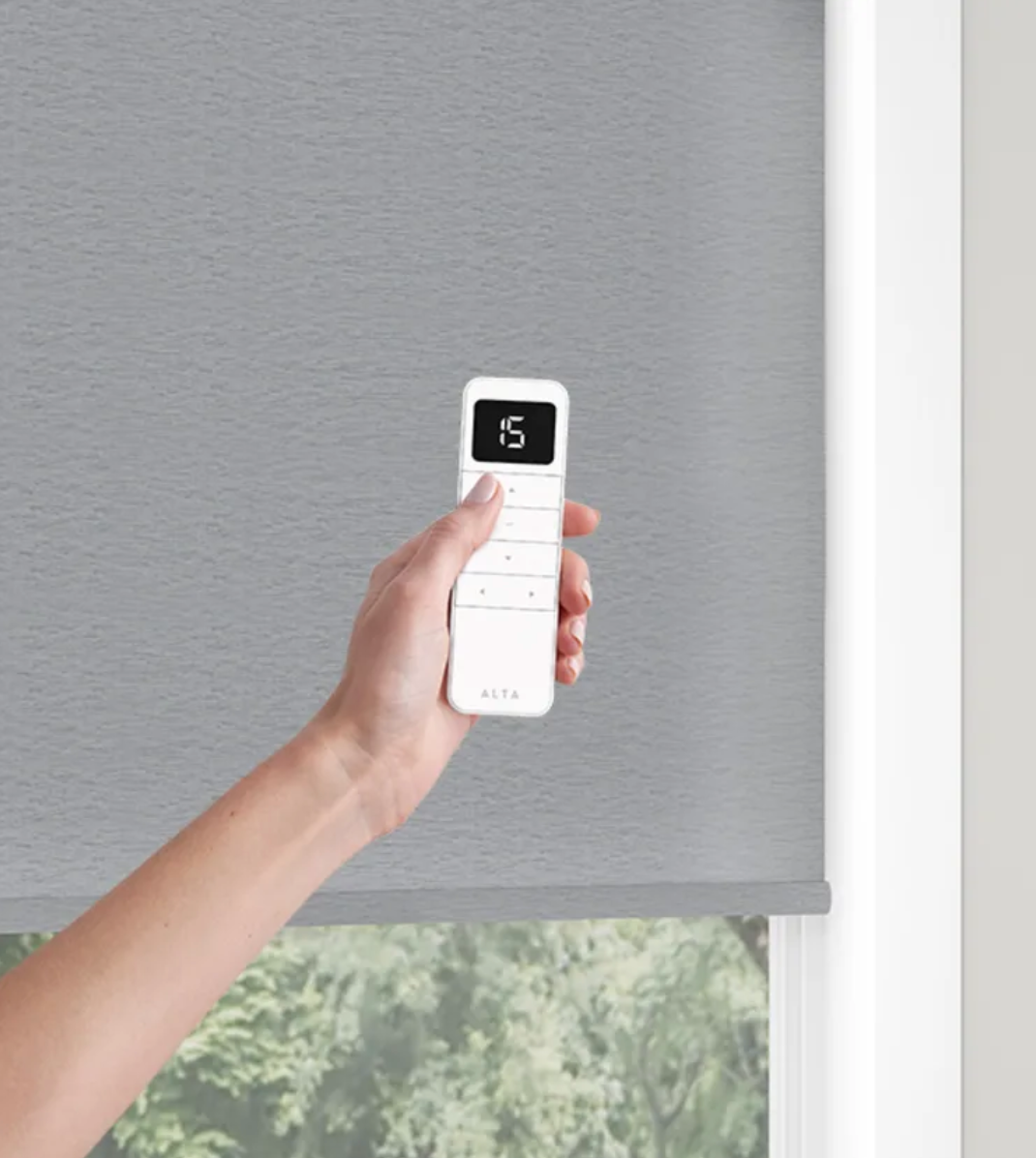 A person is holding a remote control over a window blind.