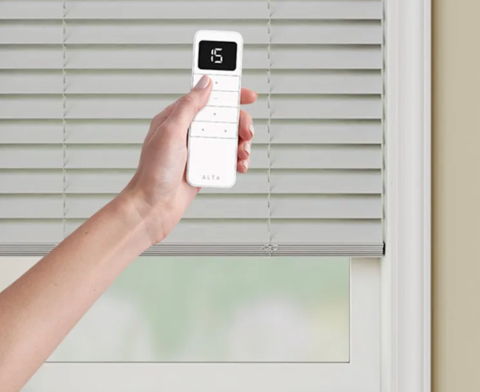 A person is holding a remote control over a window blind.