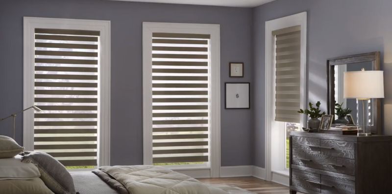 A bedroom with a bed , dresser , mirror and striped dual shades on the windows. Love is Blinds