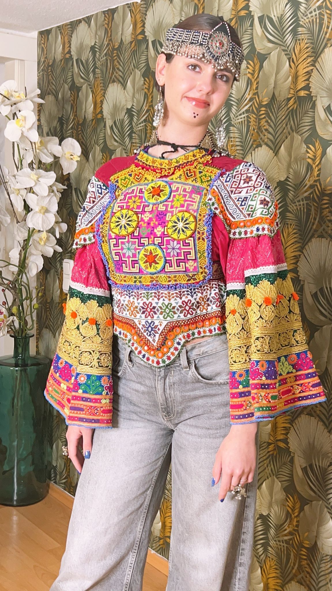 Eine Frau in einem bunten Top und Jeans steht vor einer Vase mit Blumen.