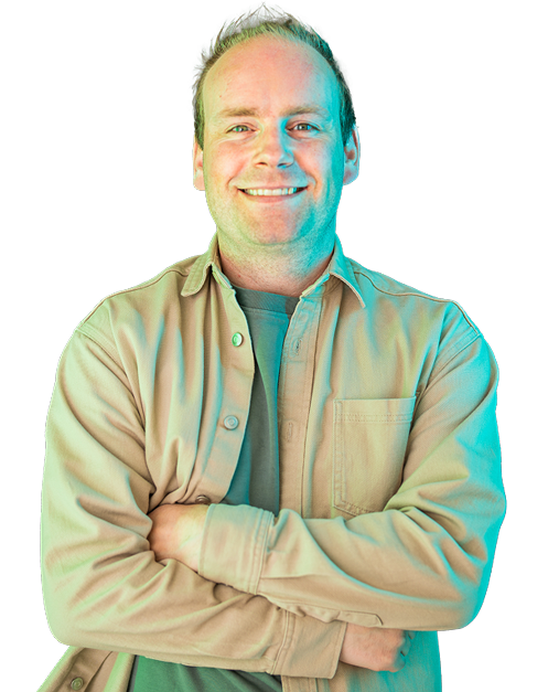 A man in a tan shirt is smiling with his arms crossed