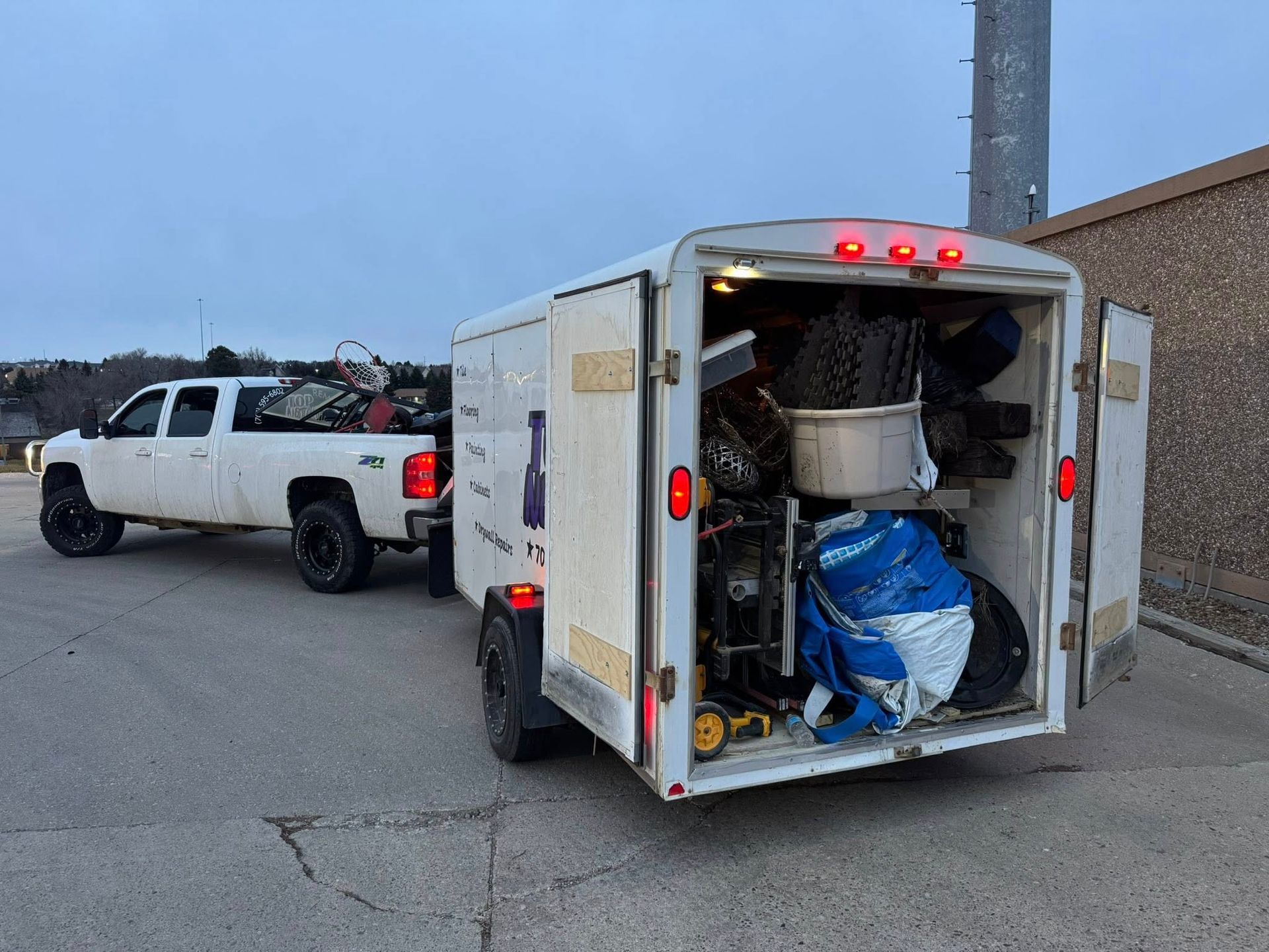 White pickup truck towing an enclosed trailer with open doors; trailer filled with assorted items.