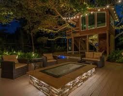 Night scene of a treehouse with outdoor seating around a lit fire pit. String lights illuminate the deck.