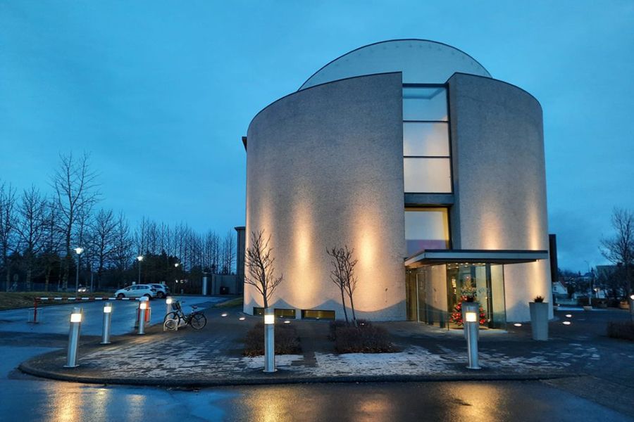Round, light-colored building with a dome roof lit up at dusk. Entrance with glass doors. Paved area with bollard lights.