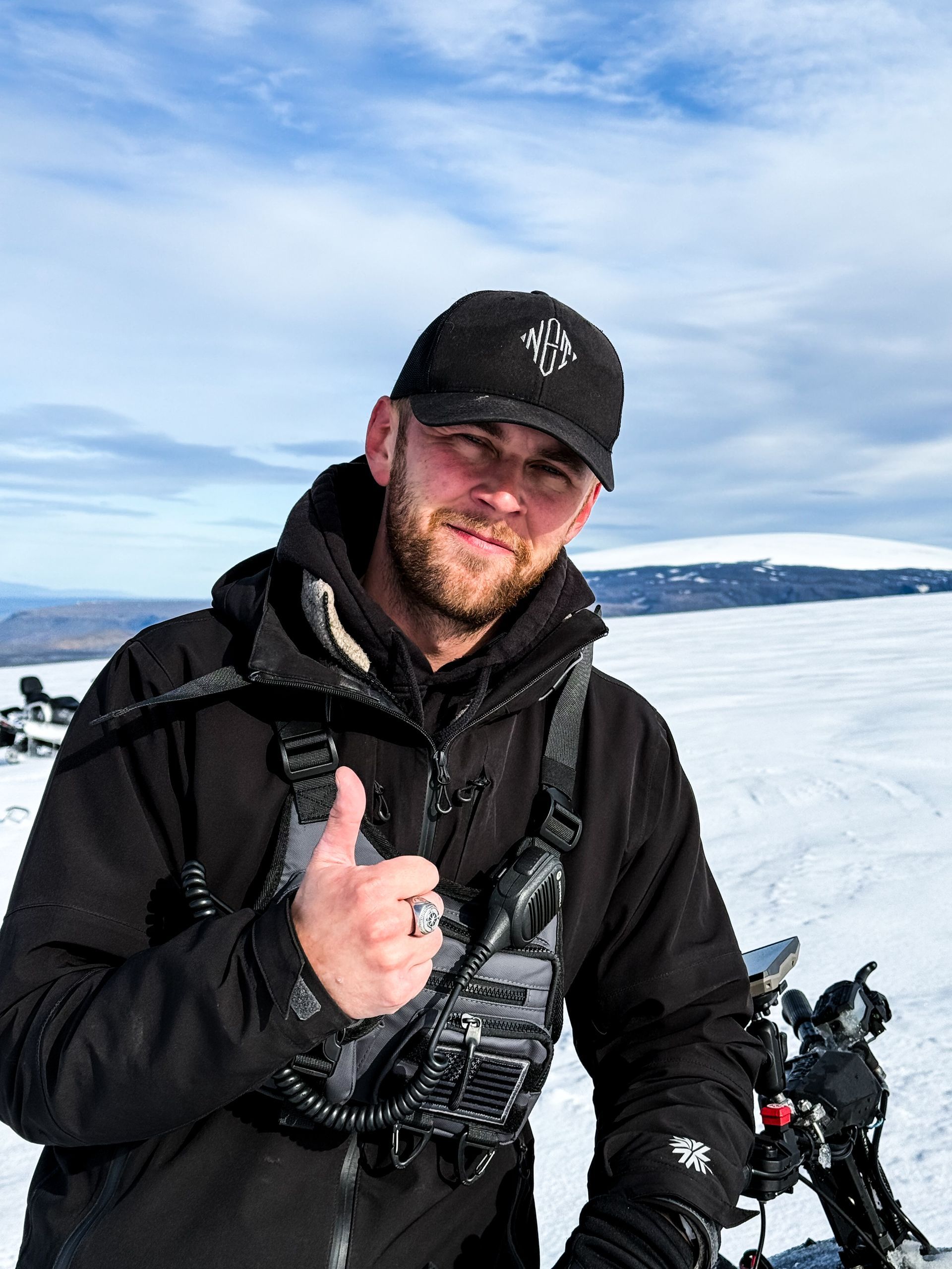 Man in black jacket and hat, thumbs up gesture, snow-covered mountain backdrop.